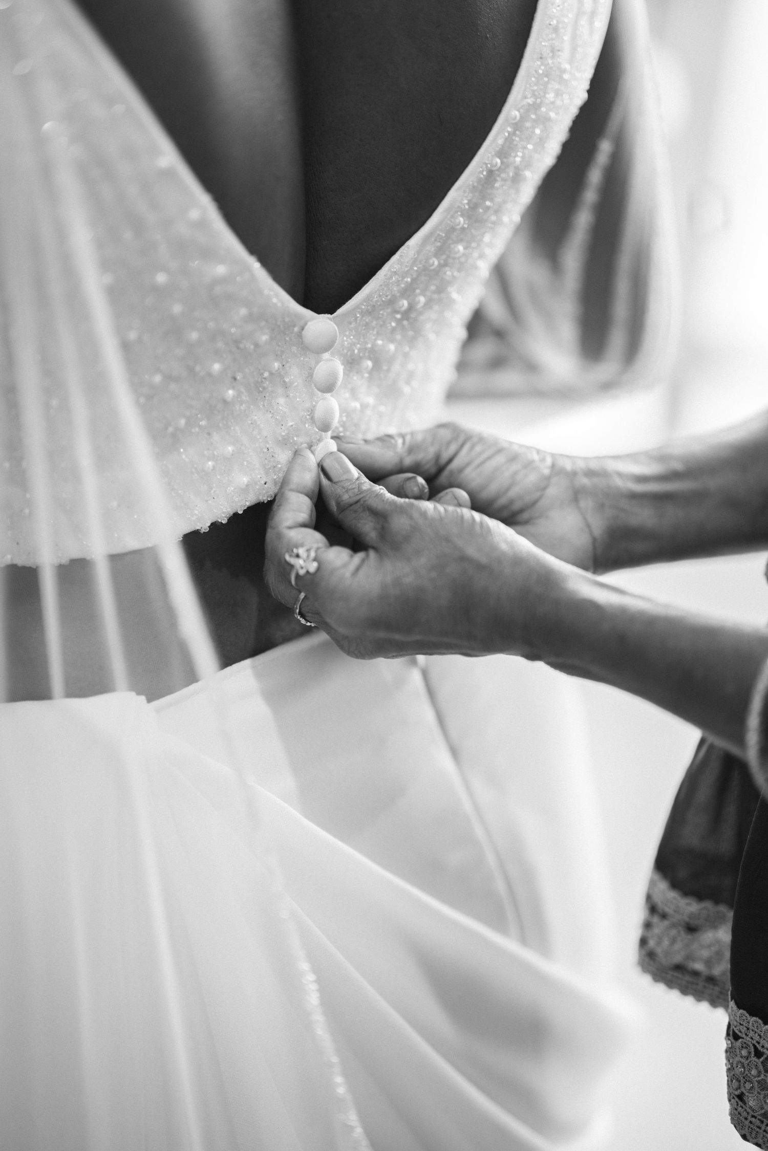 Black and white close-up of hands fastening pearl buttons on back of wedding dress.