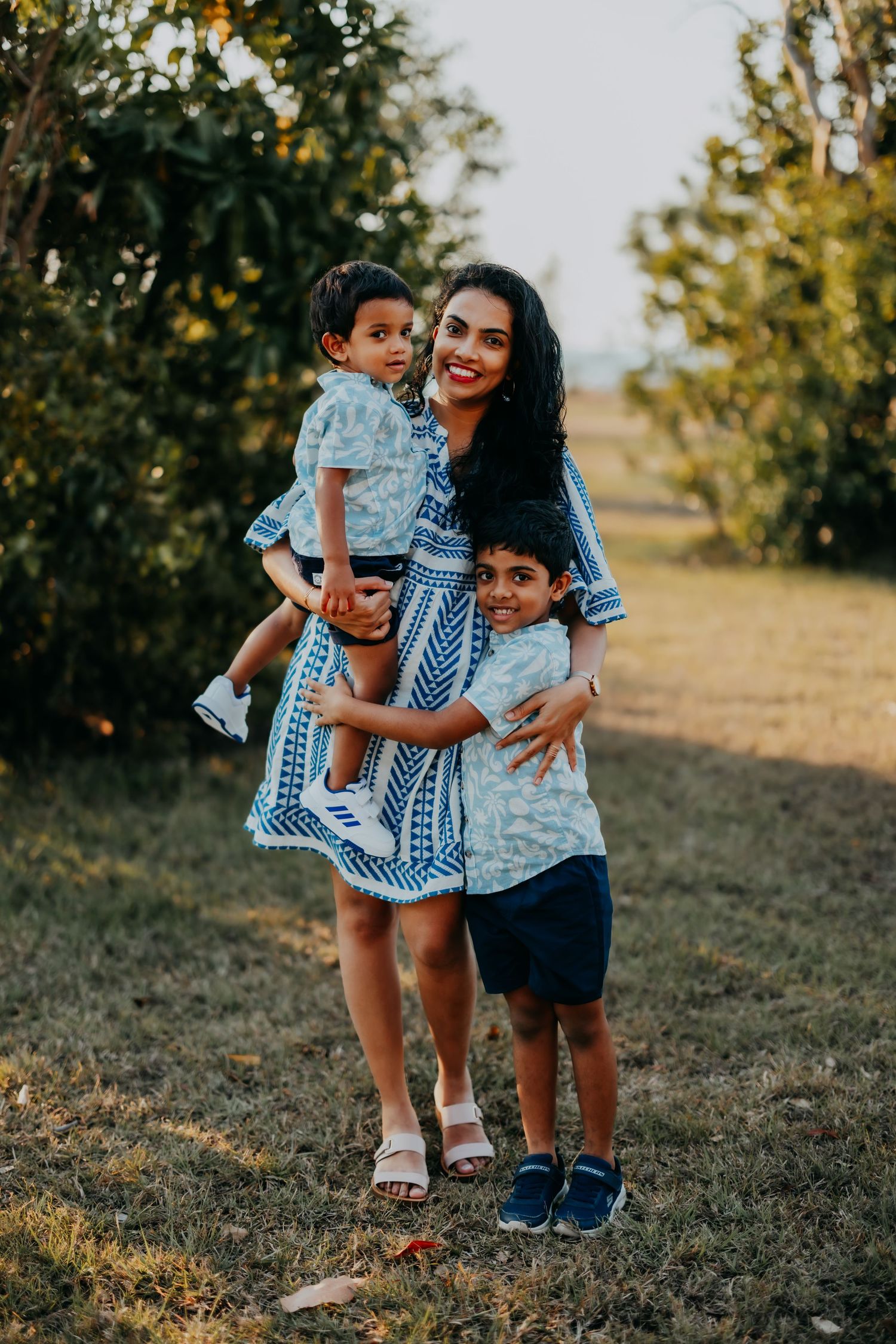 A smiling mother in a blue patterned dress holds her toddler while her older son hugs her outdoors.