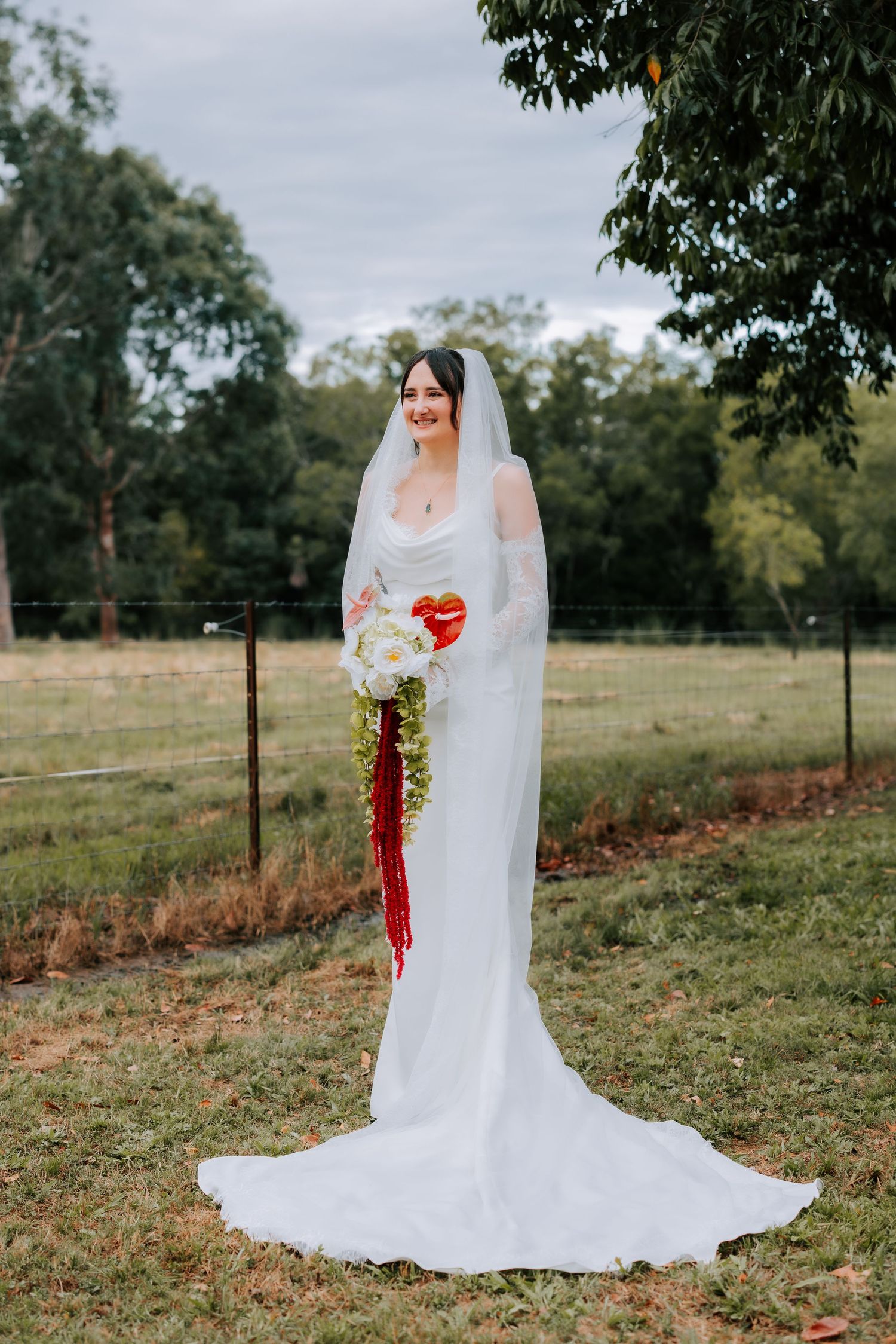 Bride in white fitted gown and veil holding red and white cascading bouquet outdoors.