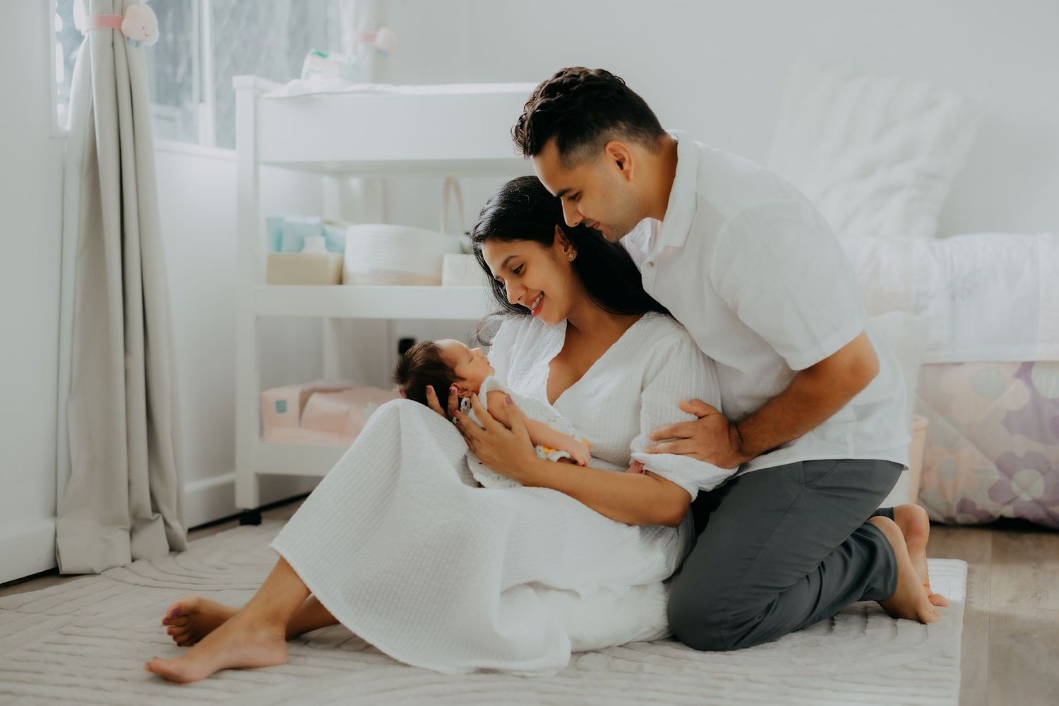 Couple sits on floor cradling newborn baby in a soft nursery room.
