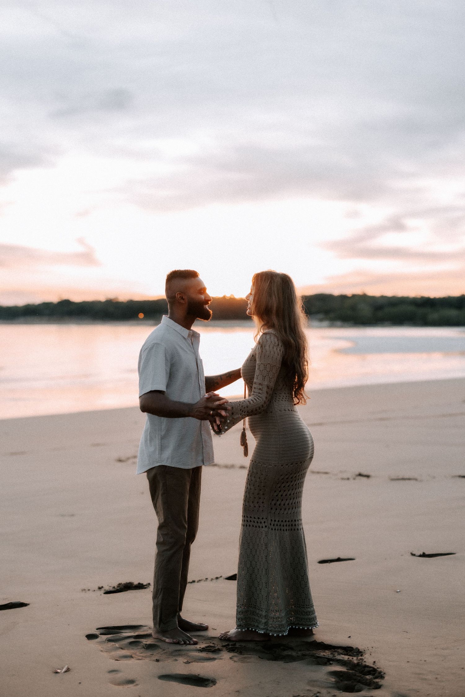 A couple holds hands facing each other on a sandy beach at sunset, with calm water and trees in the background.