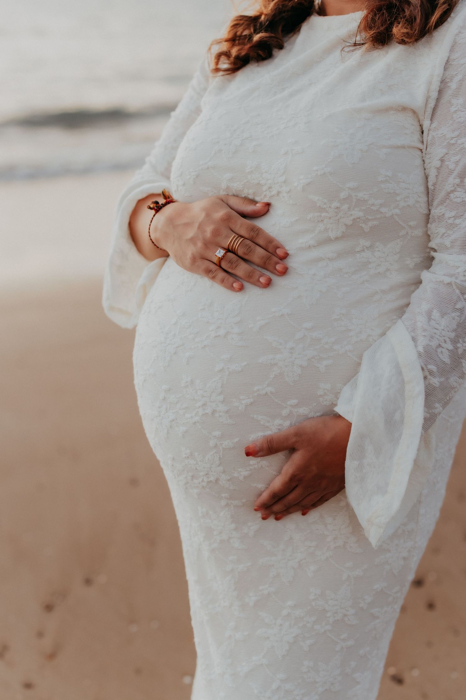 Close-up of pregnant woman in white lace dress cradling her baby bump with gold rings.