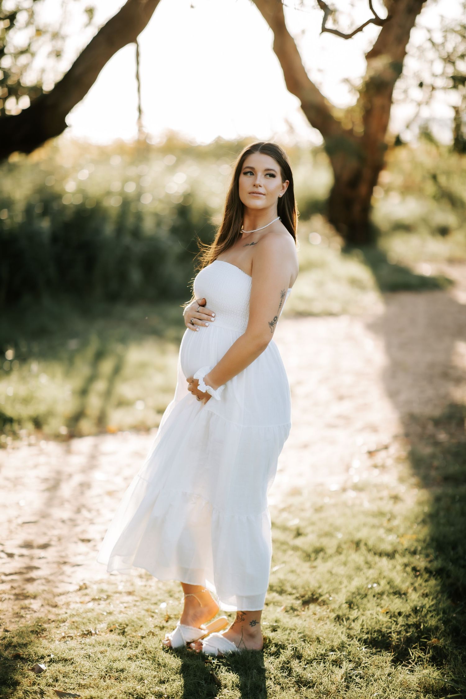 Pregnant woman in white strapless dress holding belly in sunlit garden path.