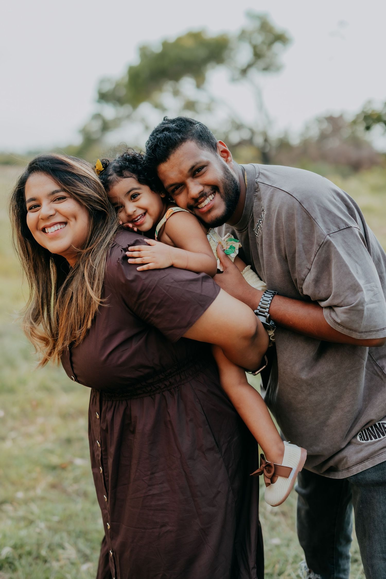 Smiling parents holding their young daughter between them in a warm outdoor family portrait in a grassy field.