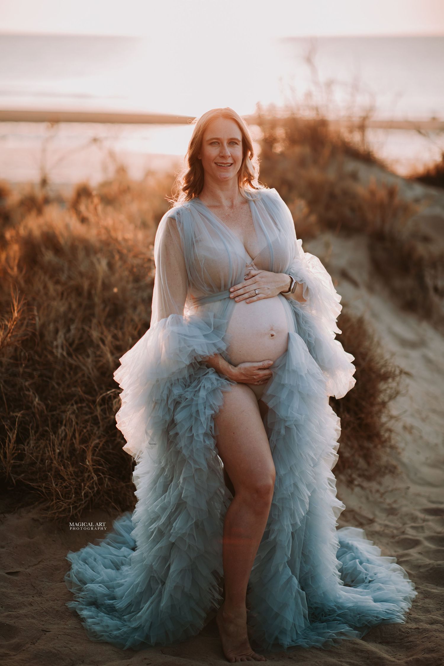 Smiling pregnant woman in sheer light blue ruffled maternity gown posing on sandy beach at sunset.