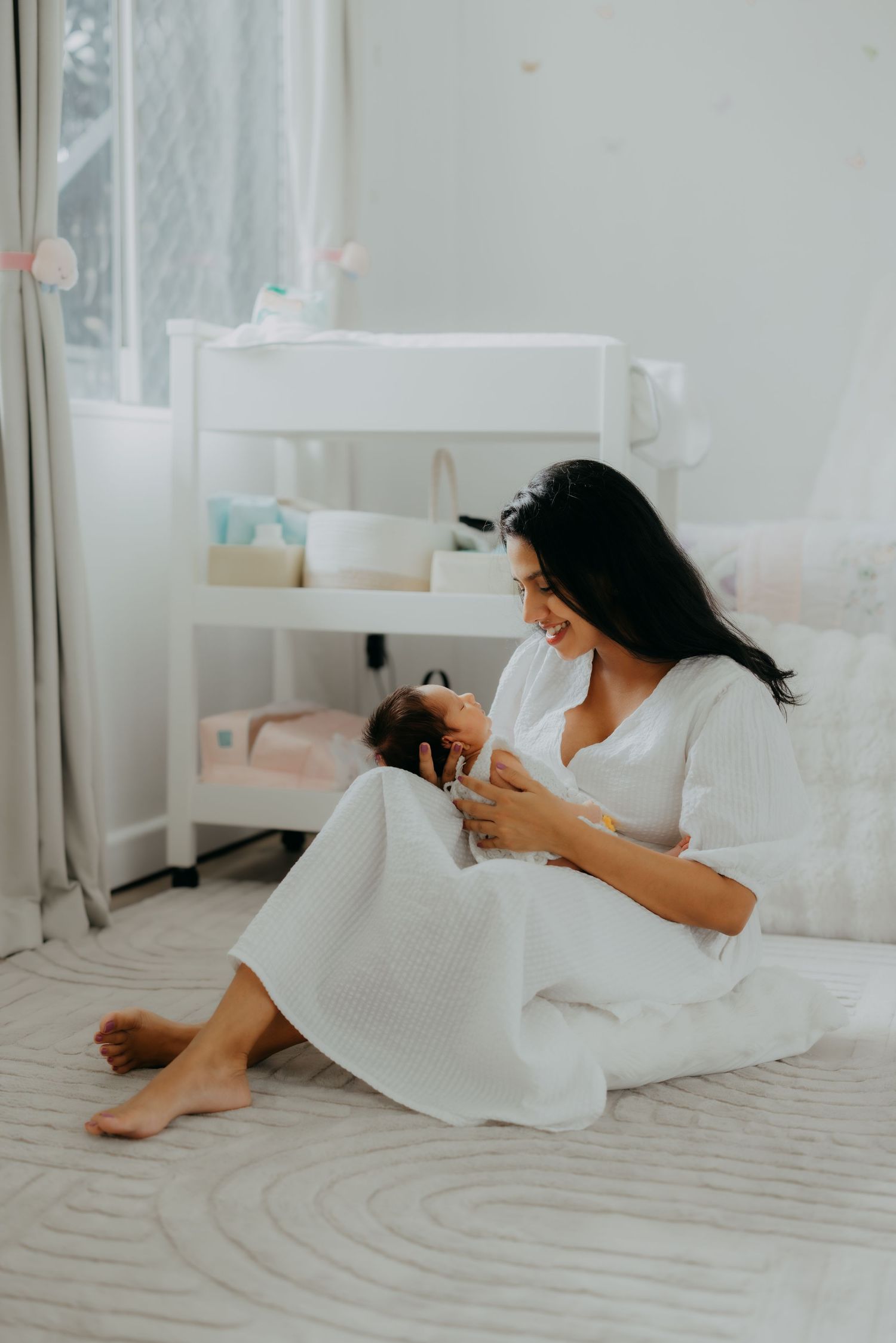 New mother in white dress sits on floor smiling while feeding her newborn baby in a nursery room.