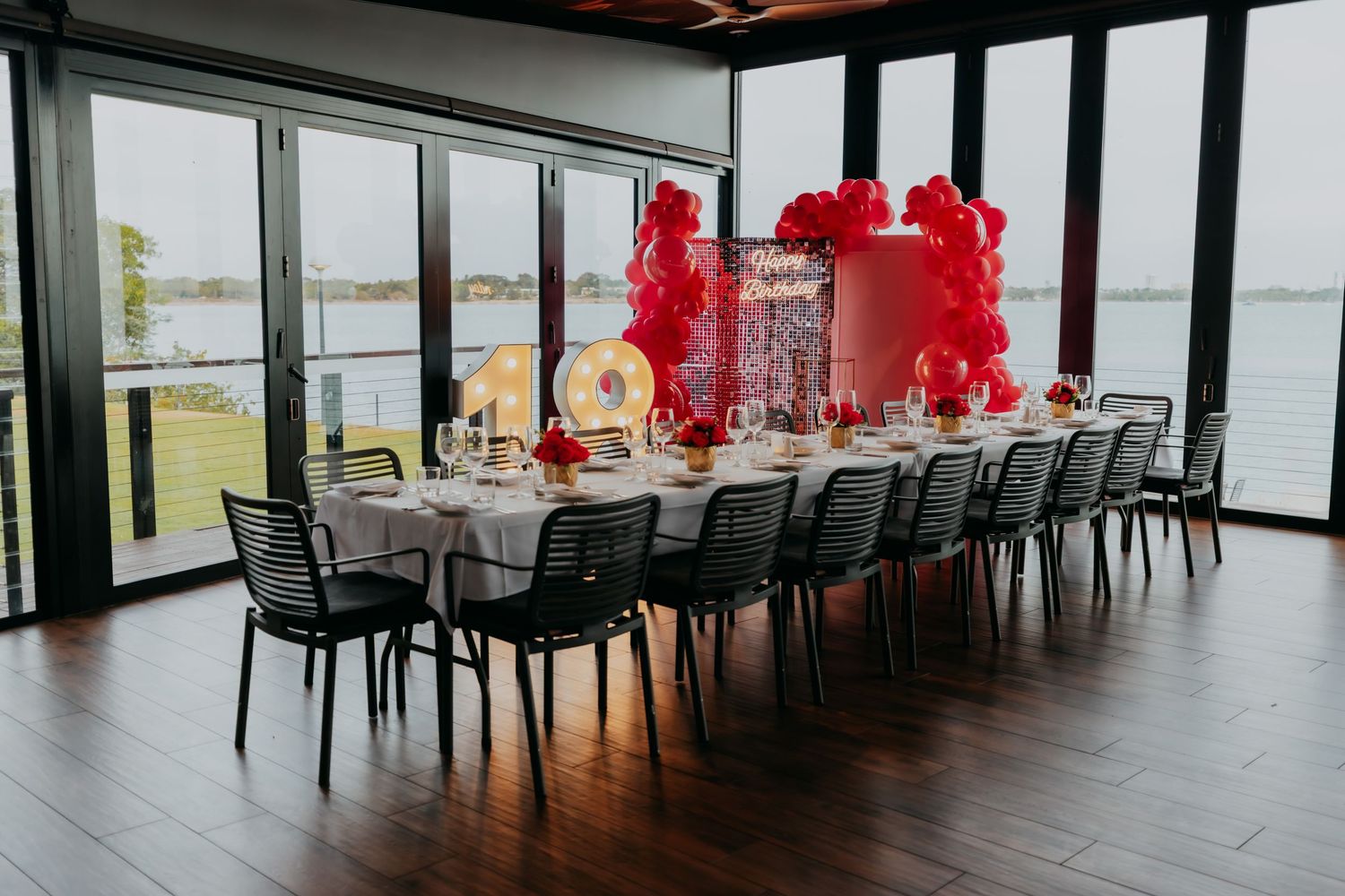 Elegant waterfront event dining room with long table, black chairs, red balloon arch centerpiece, and panoramic water views.