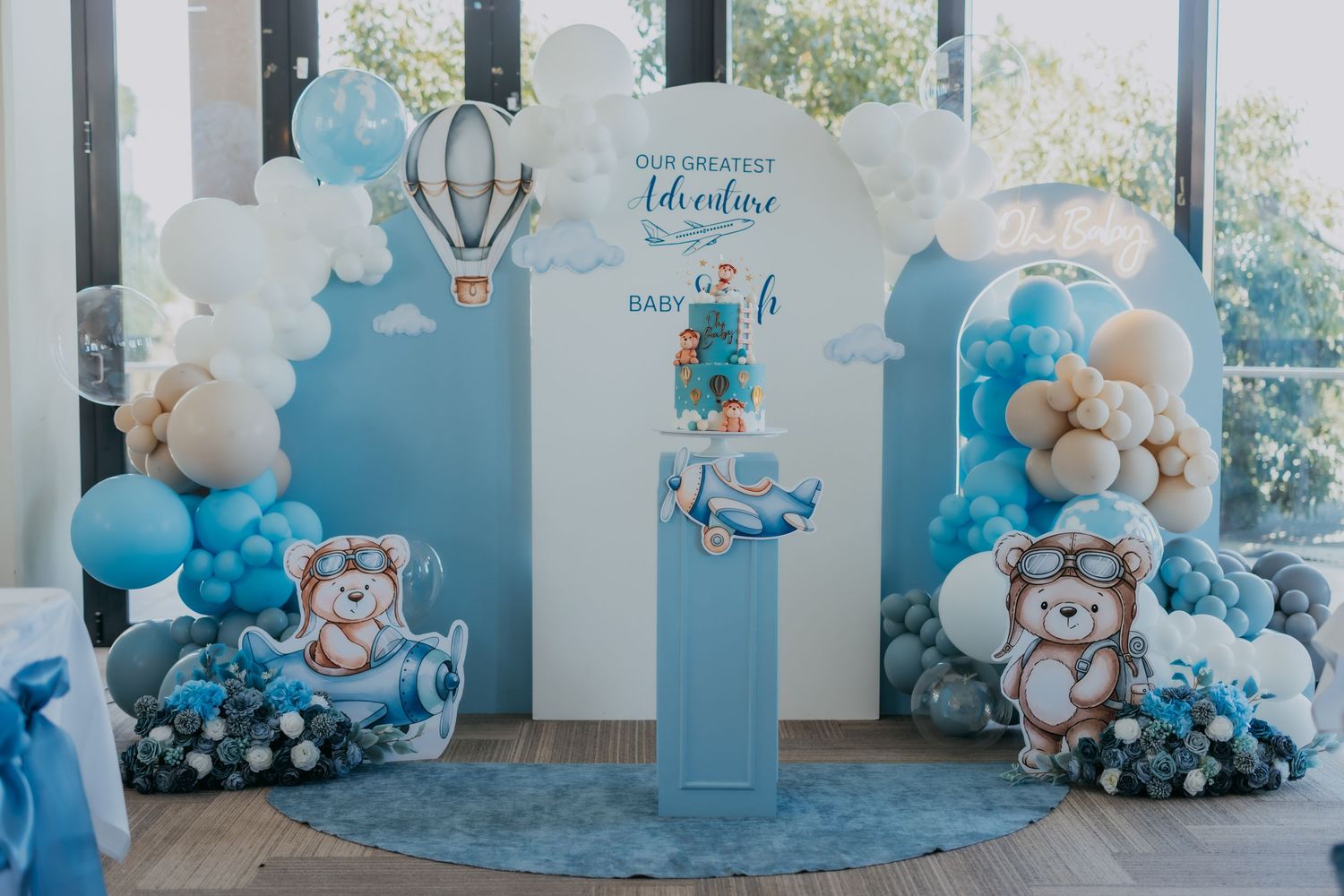 Blue teddy bear adventure baby shower decoration setup with balloon garland, hot air balloon, and bear character props.
