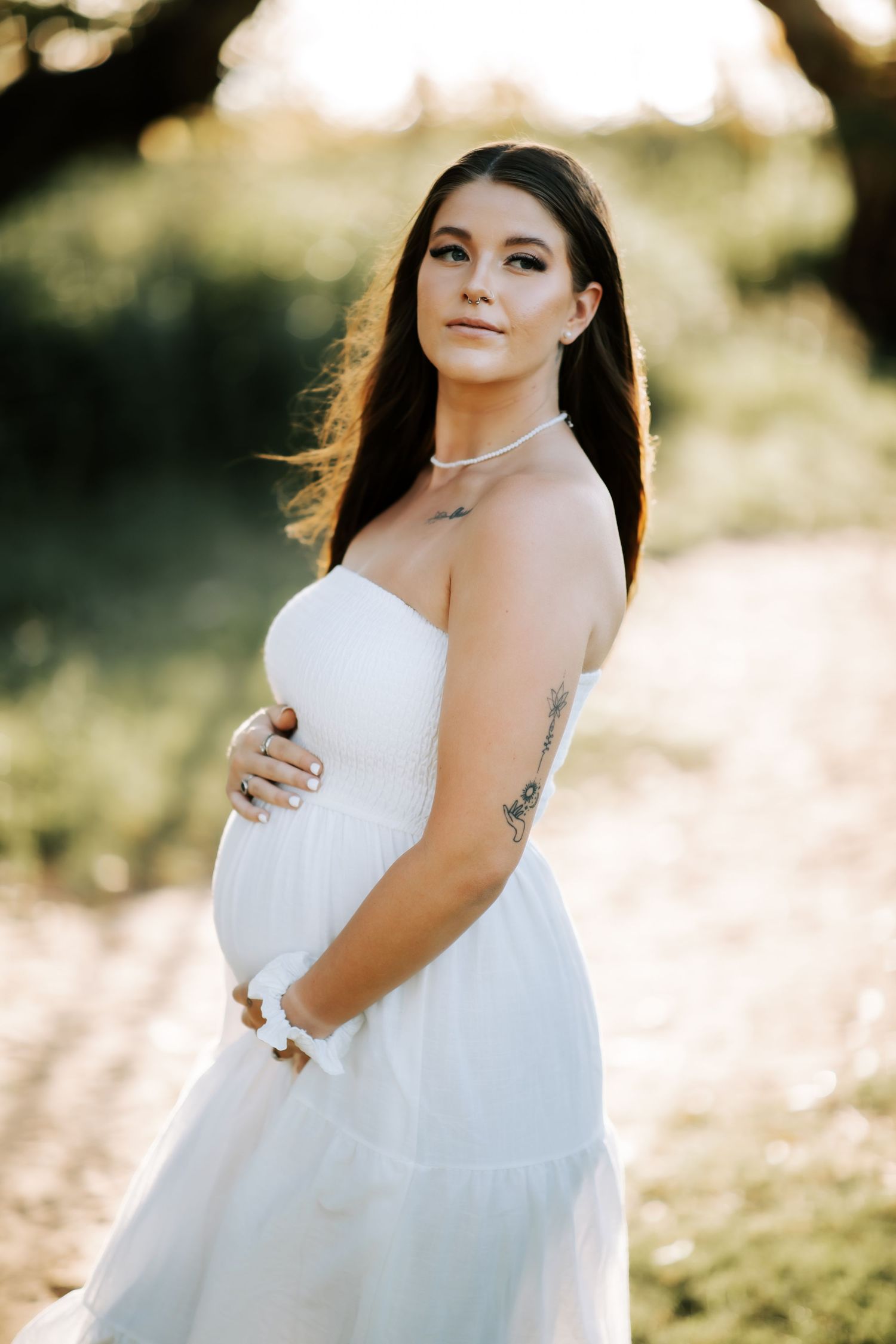 Pregnant woman in white strapless dress with tattoo poses outdoors in golden sunset light.
