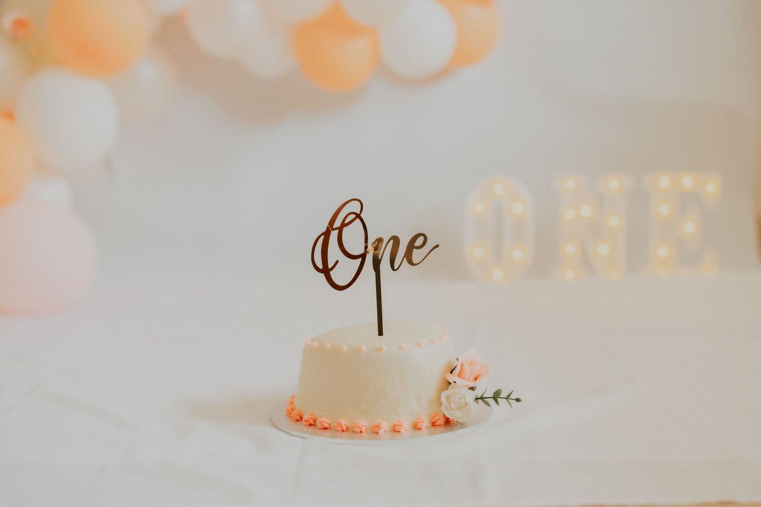 Elegant first birthday smash cake with gold One topper, pink floral details, and peach balloon garland backdrop.