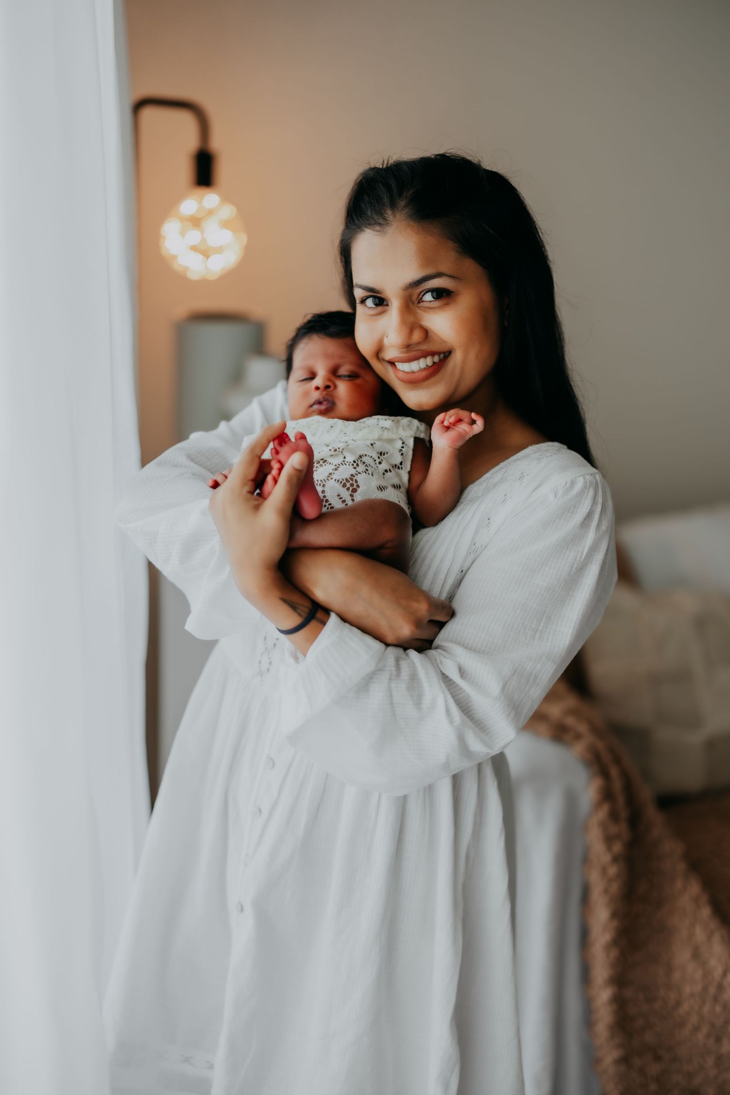 Smiling mother in white dress holds newborn baby near window light.