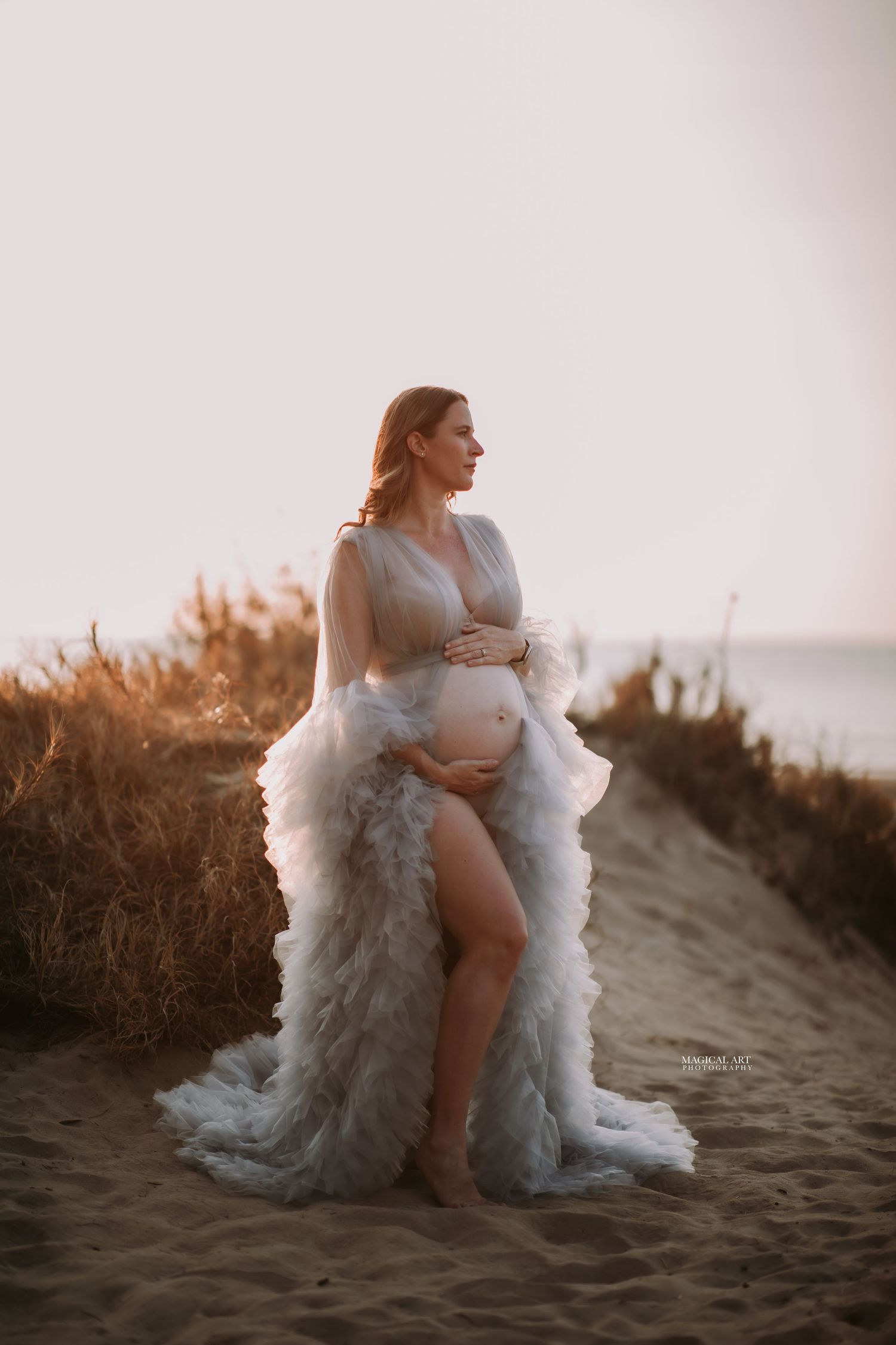 Pregnant woman in elegant white ruffled maternity robe standing on sandy beach path at dusk.