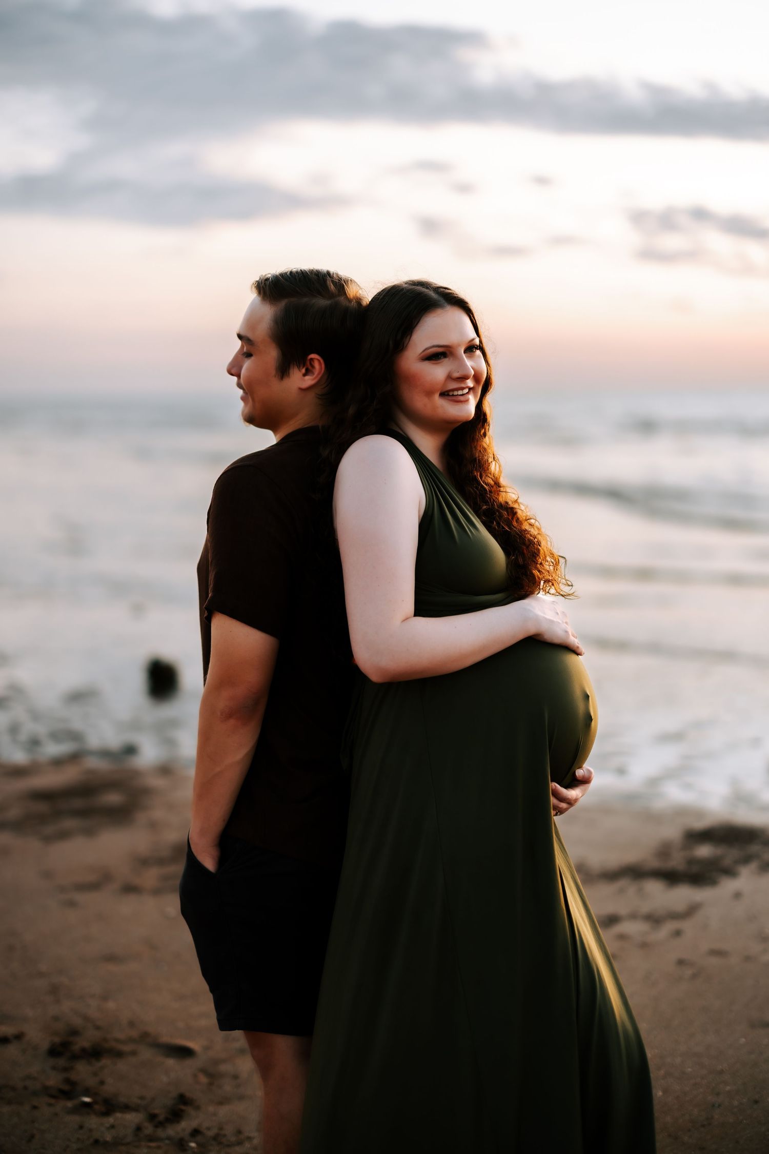 Pregnant woman in green dress smiling on beach at sunset while partner stands back-to-back.