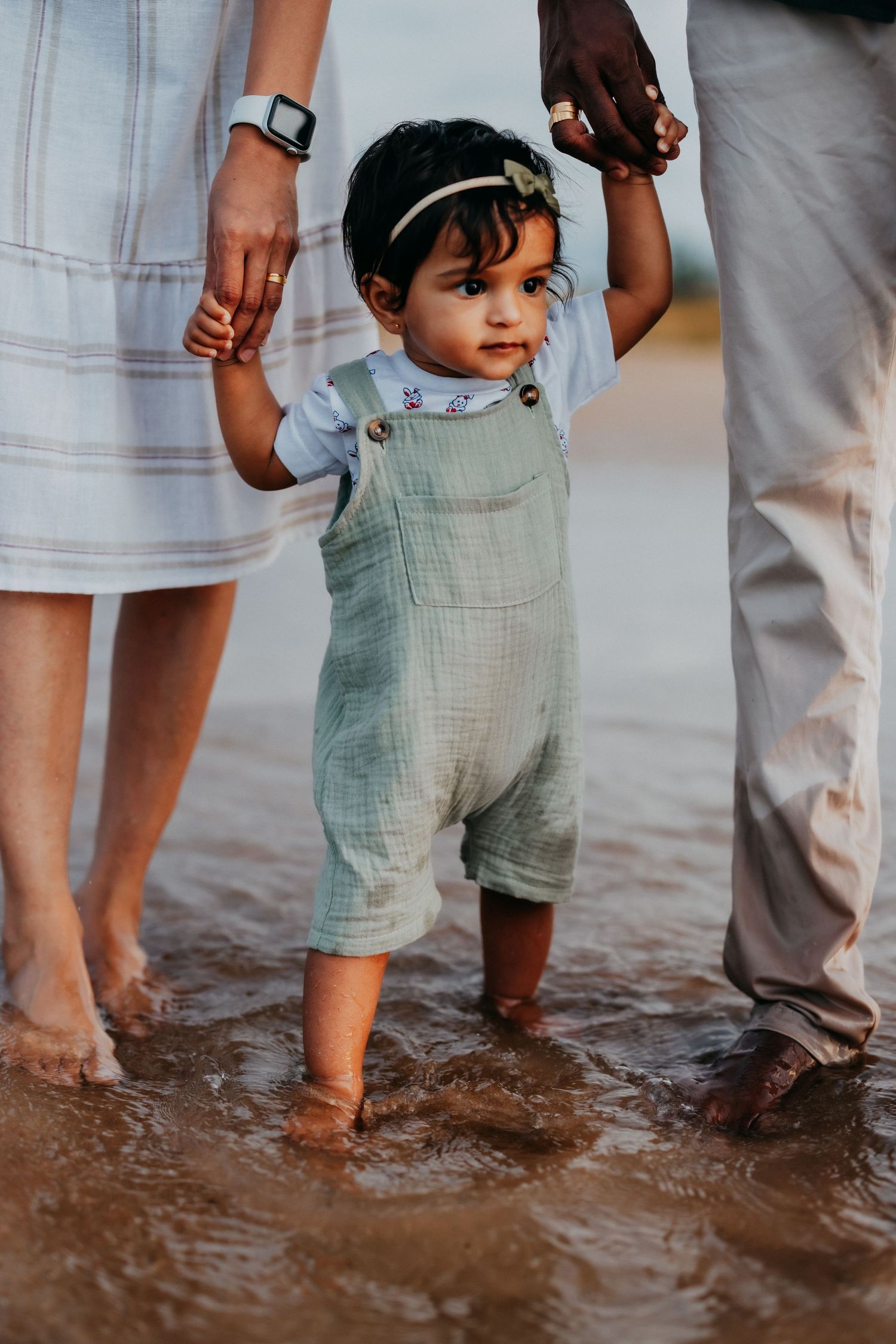 An adorable baby girl in green overalls takes steps in shallow water, held by both parents.