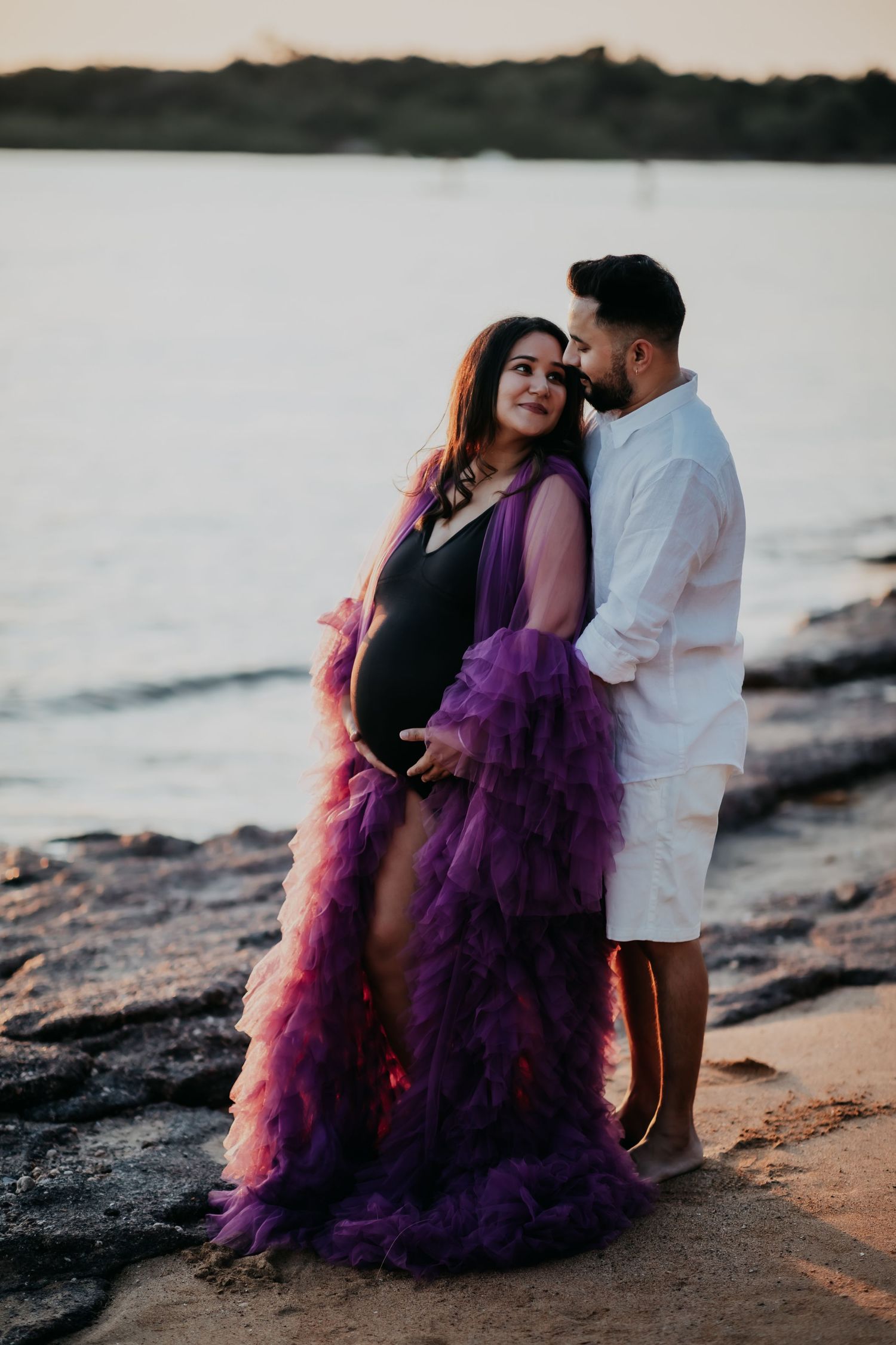 Pregnant couple embracing on rocky beach at sunset, woman in purple tulle maternity gown.