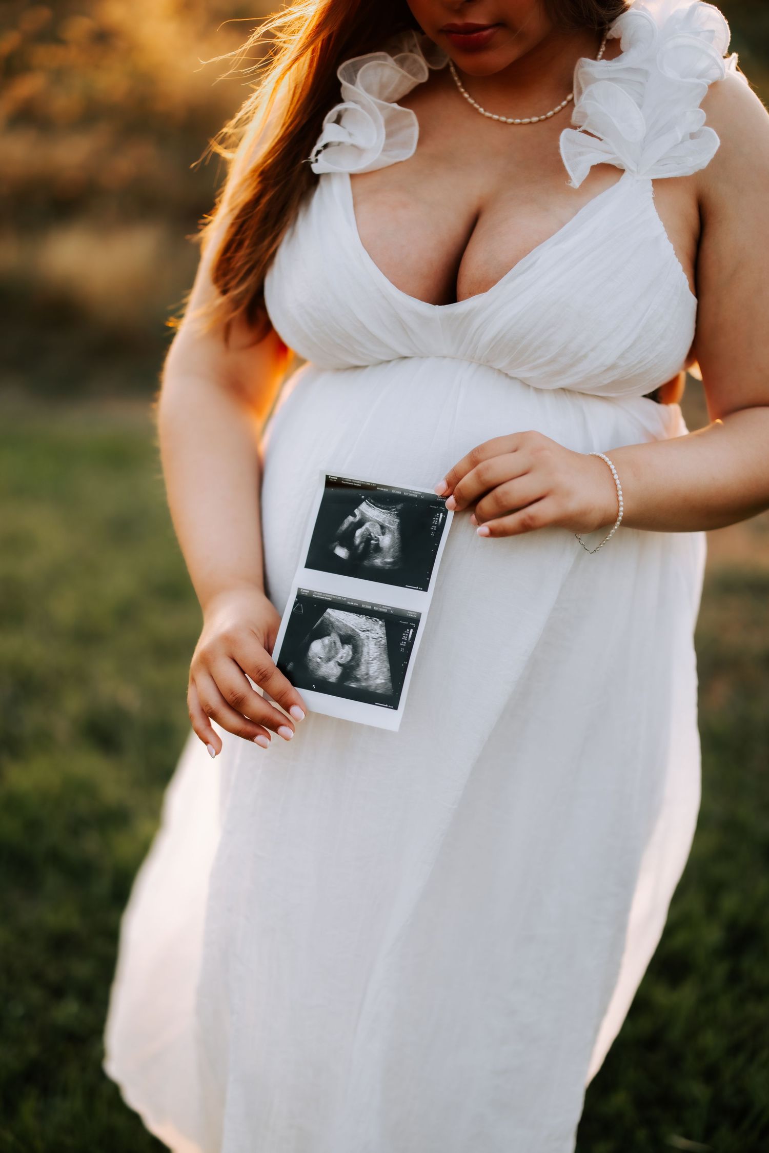 Pregnant woman in white dress holding ultrasound photos against baby bump in outdoor setting.