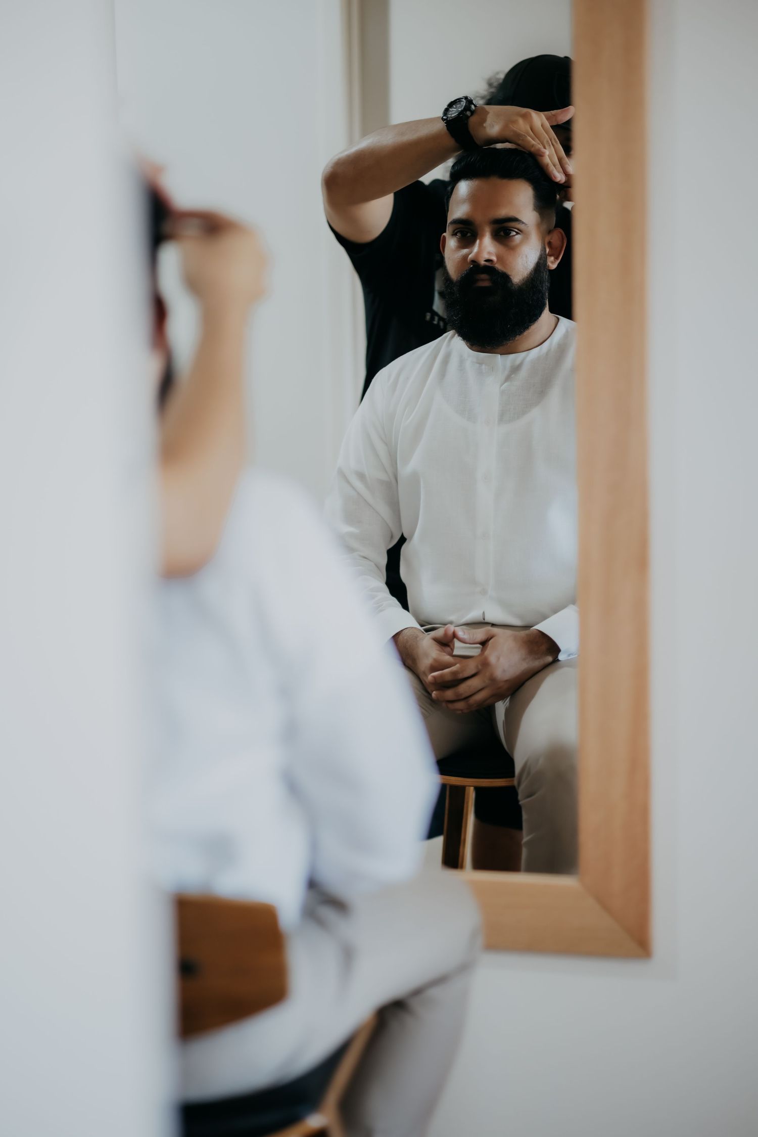 Groom with beard getting hair styled by hairdresser reflected in large mirror.