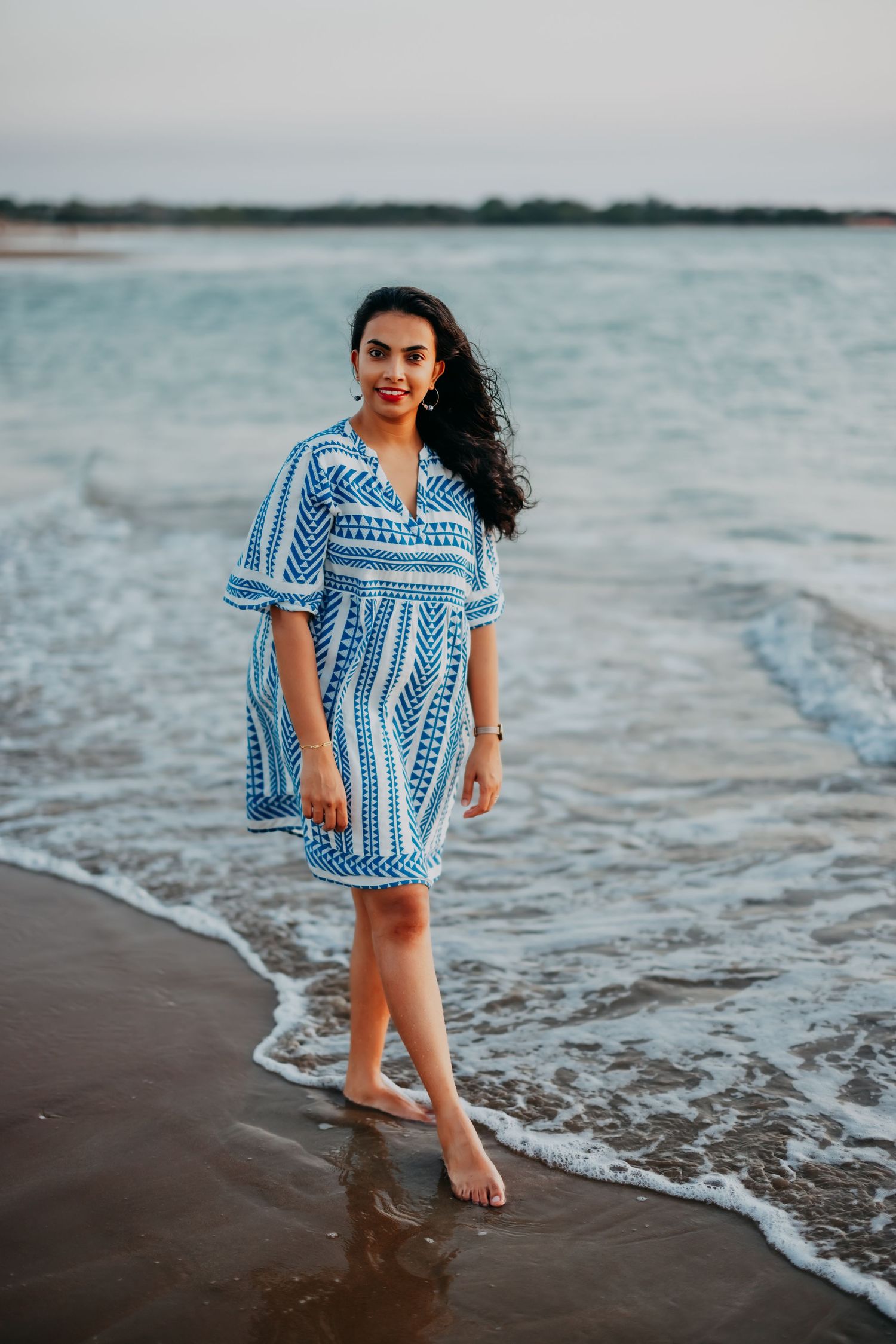 Young woman in blue and white patterned dress standing barefoot at the ocean's edge on a sandy beach.