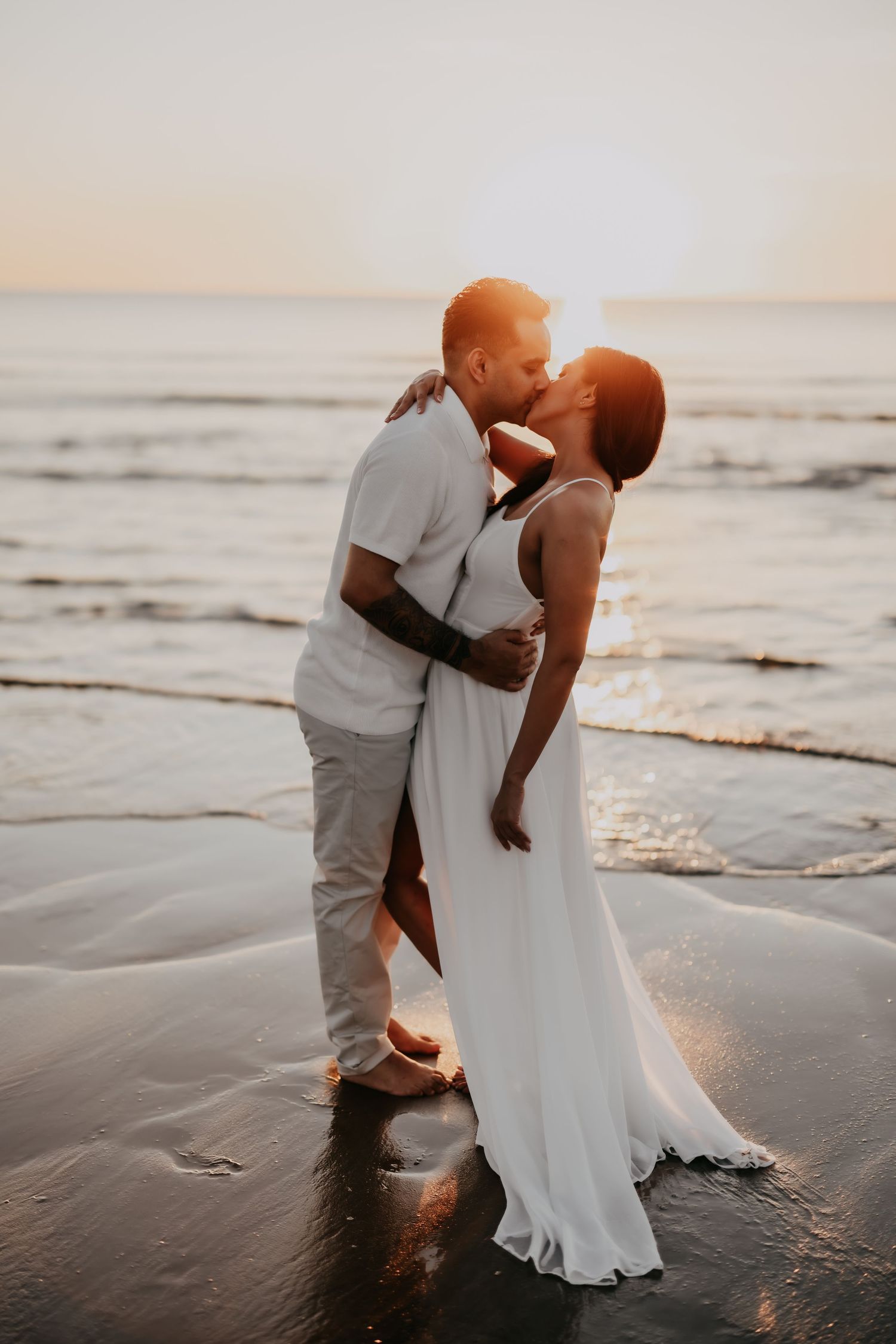 Couple sharing a passionate kiss at sunset on a wet beach with golden ocean reflections around them.