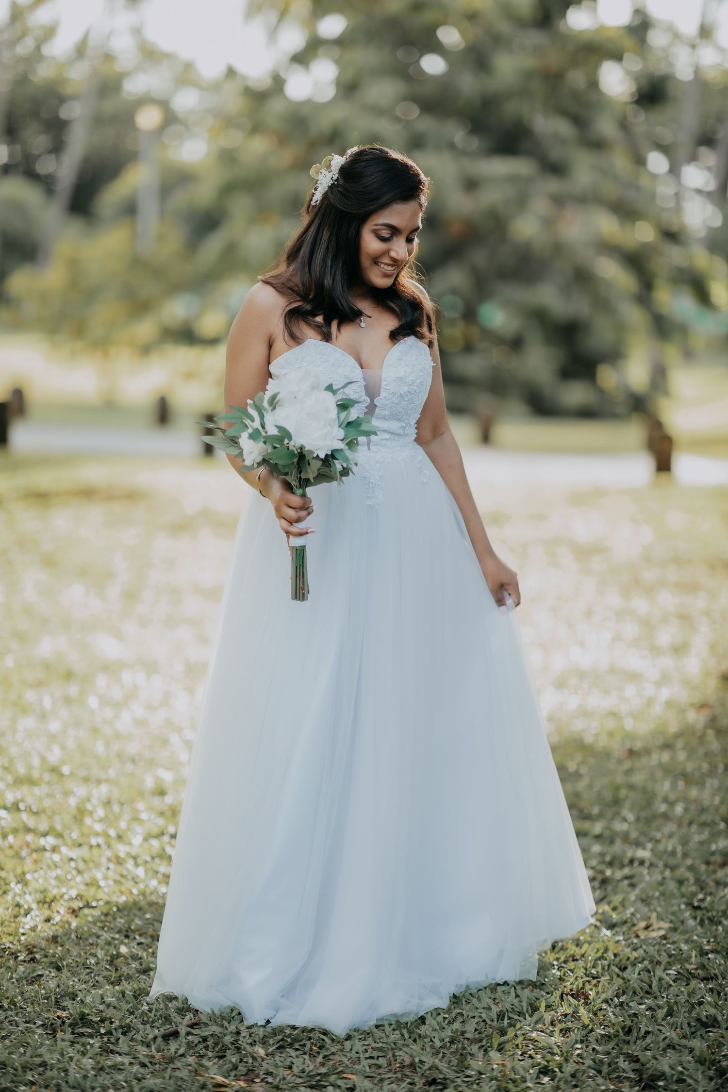 Stunning bride in a white lace tulle gown holding a white bouquet in a lush green garden.