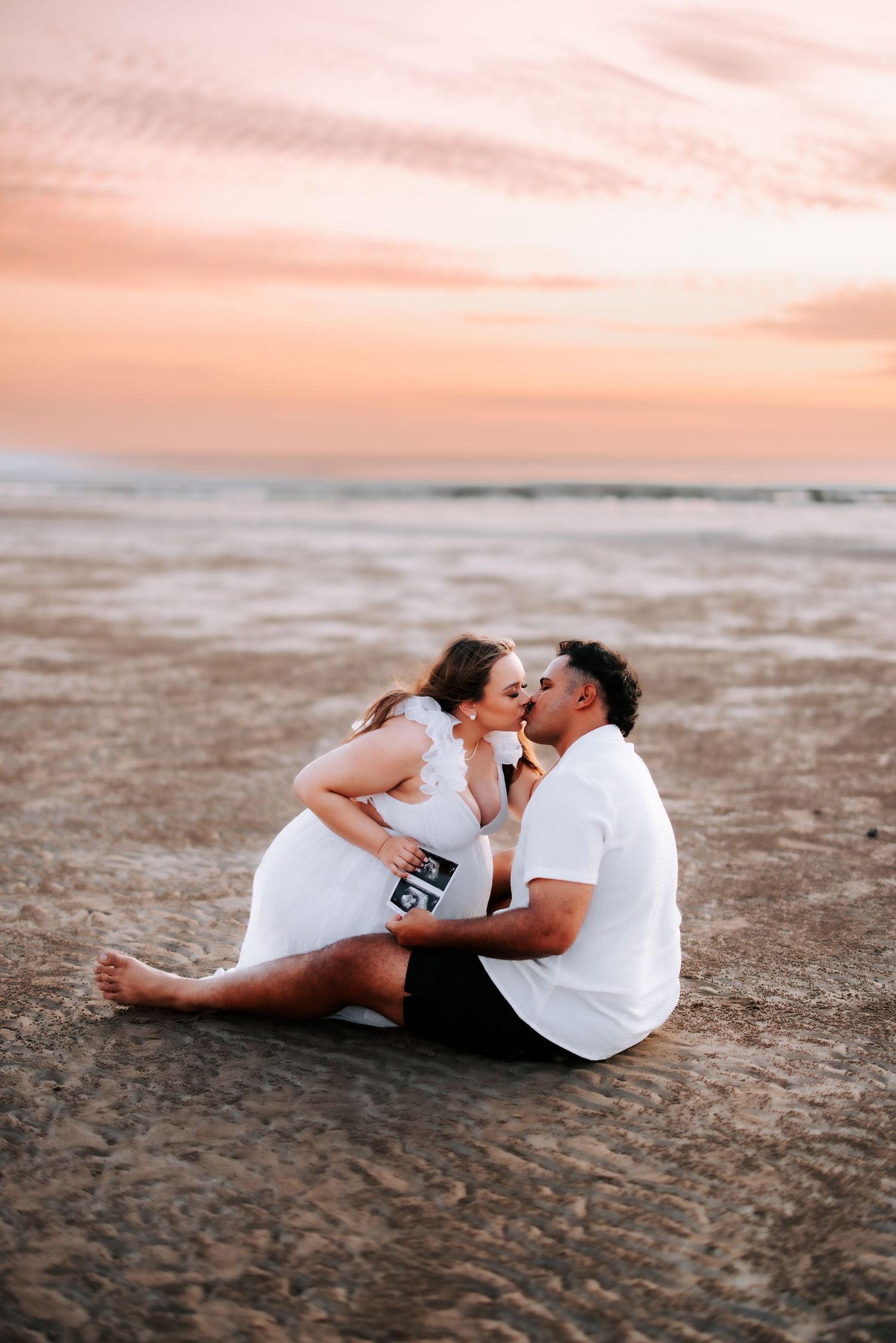 Pregnant couple kissing while sitting on beach at pink sunset, holding ultrasound photo.