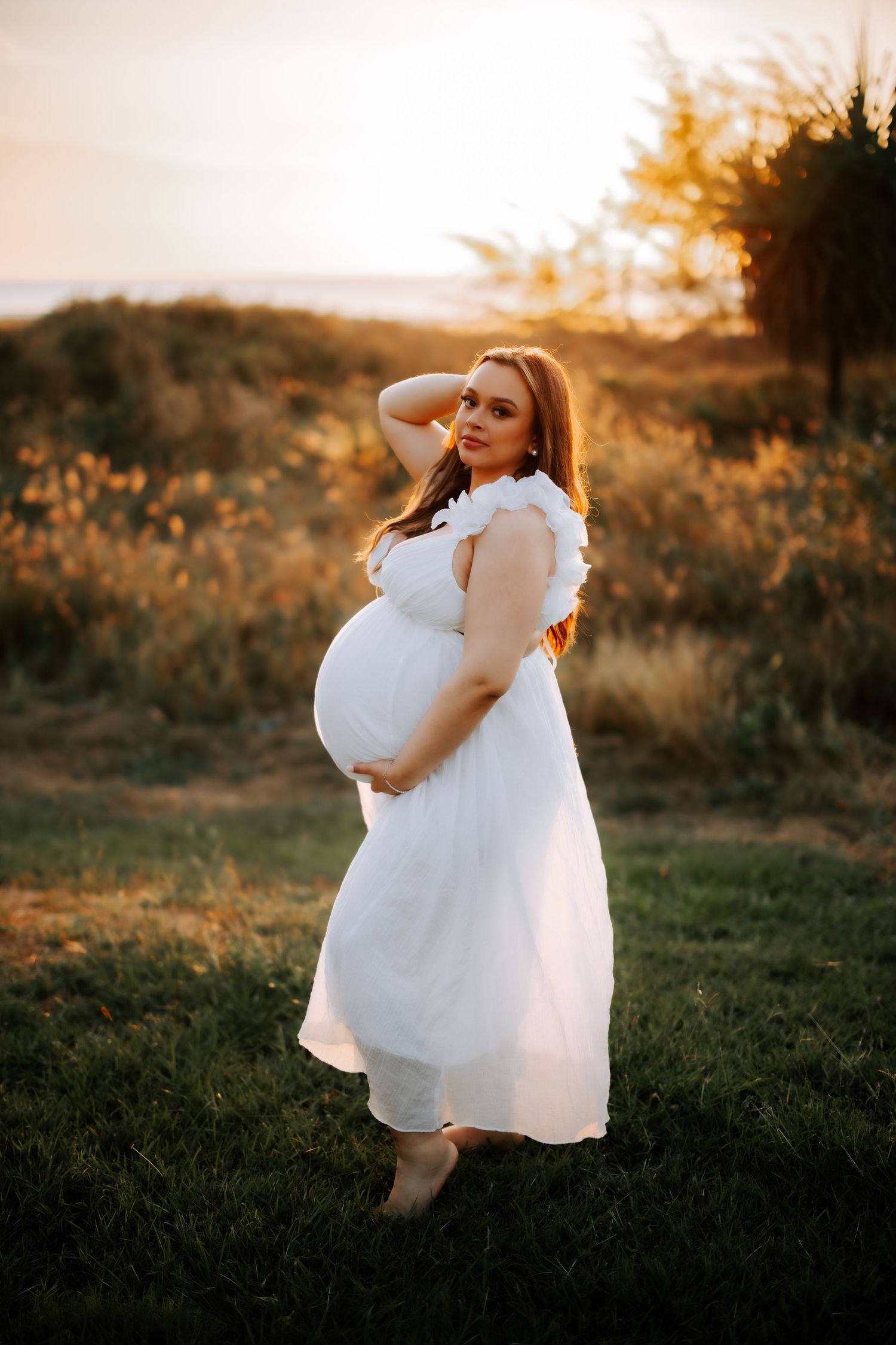 Pregnant woman in flowing white dress cradling baby bump during golden sunset in a scenic outdoor field.