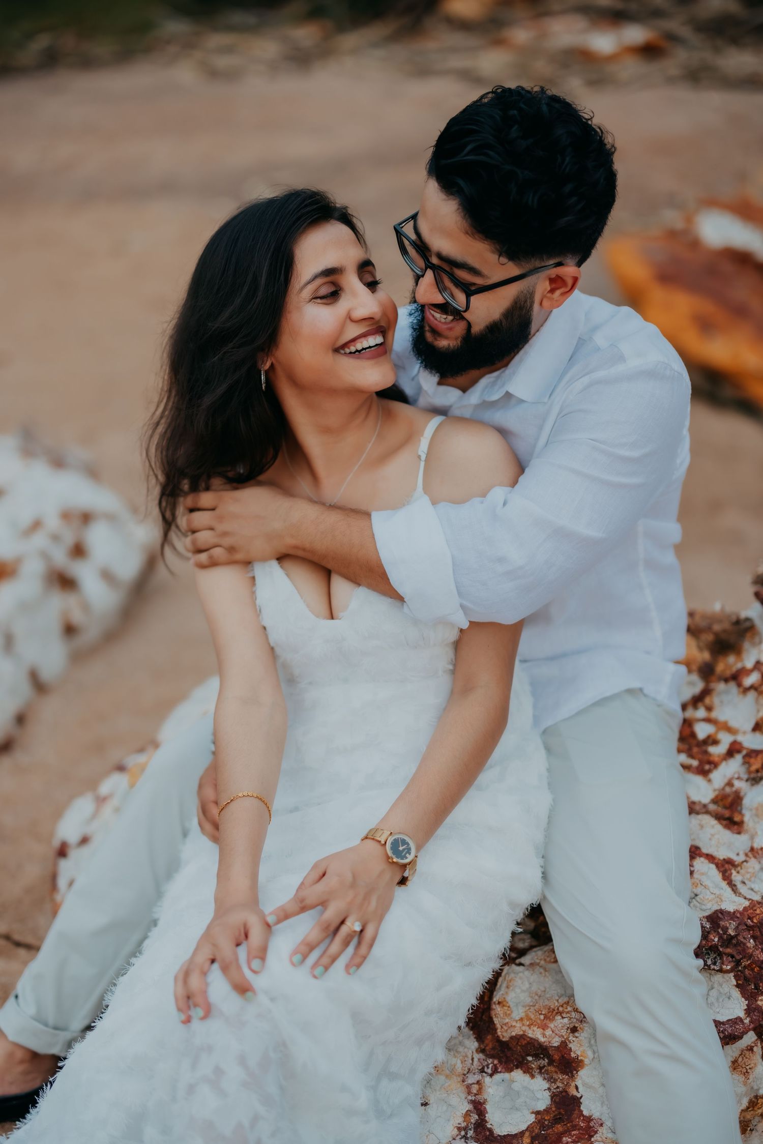 Couple in wedding attire sharing romantic moment sitting on rocks during outdoor engagement shoot.