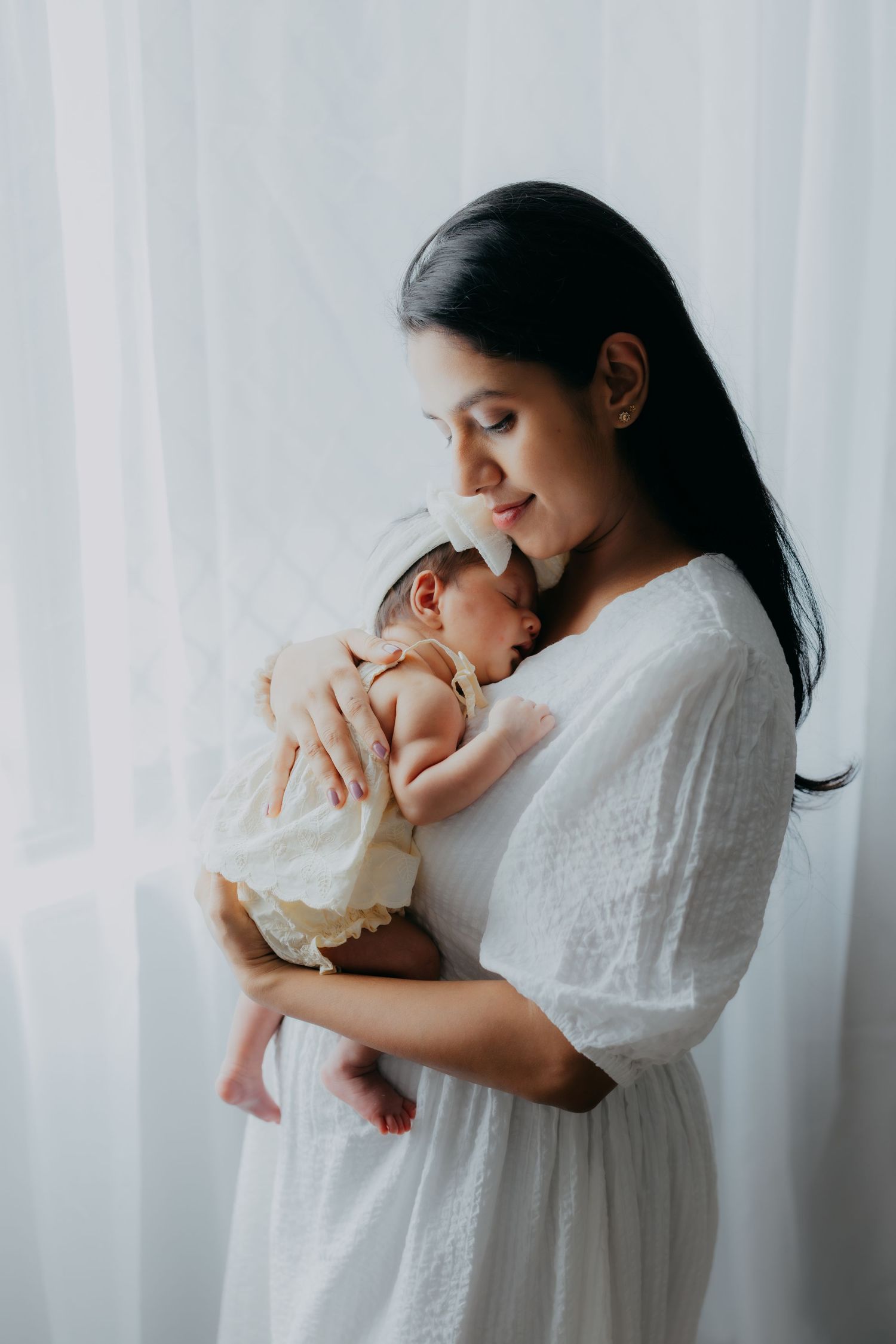 Mother in white dress tenderly holding and cuddling her newborn baby near sheer curtains.