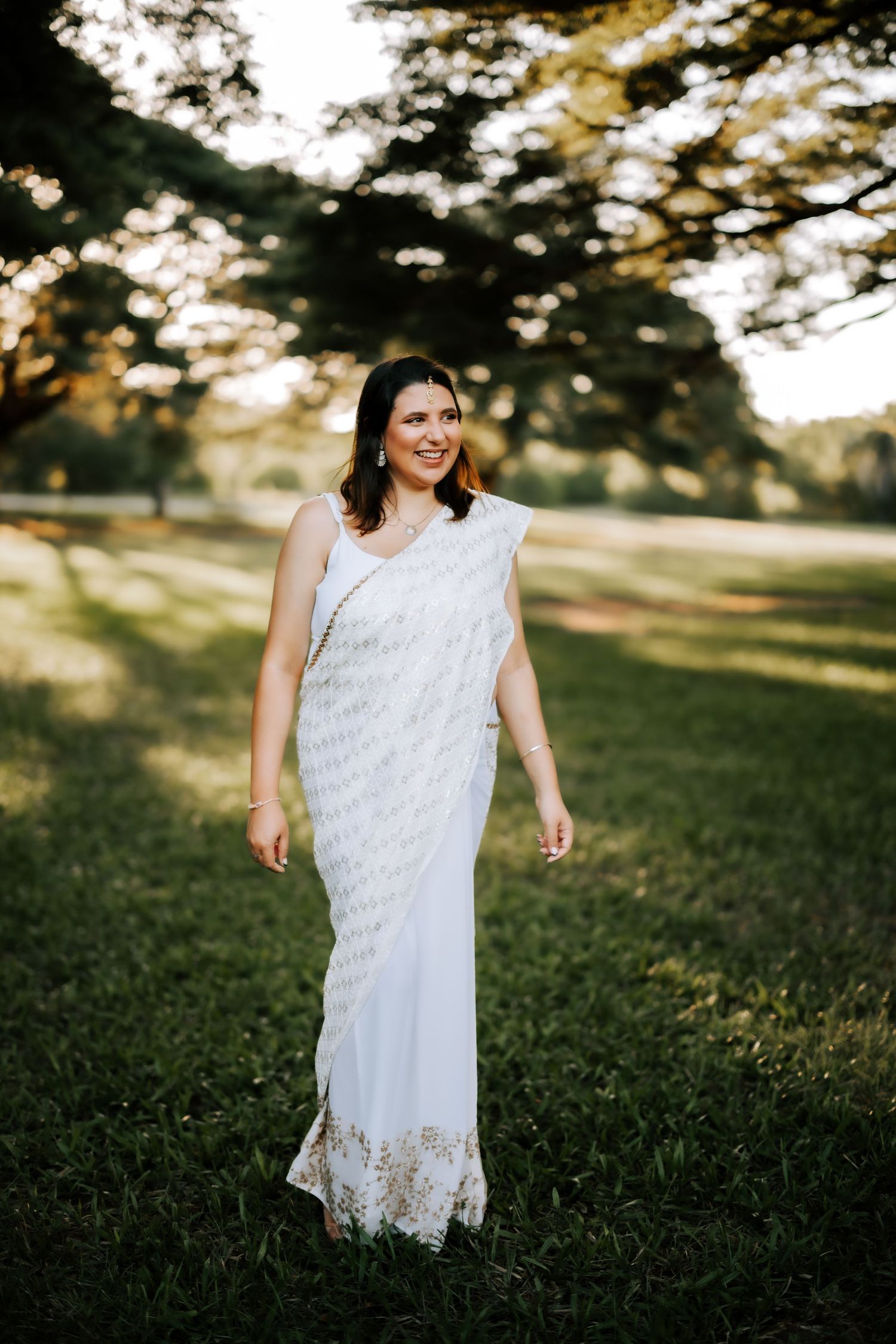 Elegant woman in white and gold embroidered saree walks through sunlit green park at golden hour.