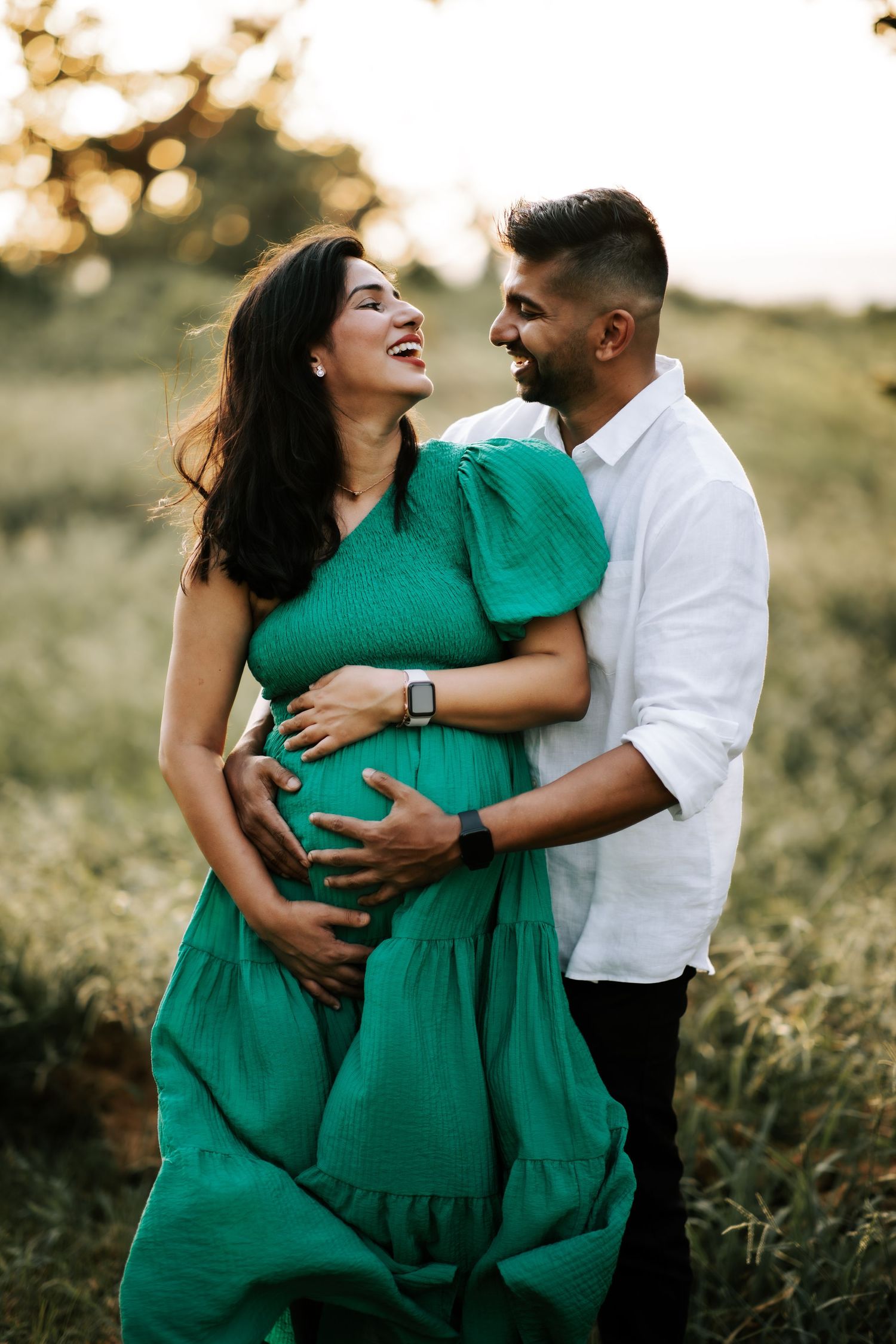 Laughing pregnant woman in green dress embraced by man in white shirt in sunlit field.