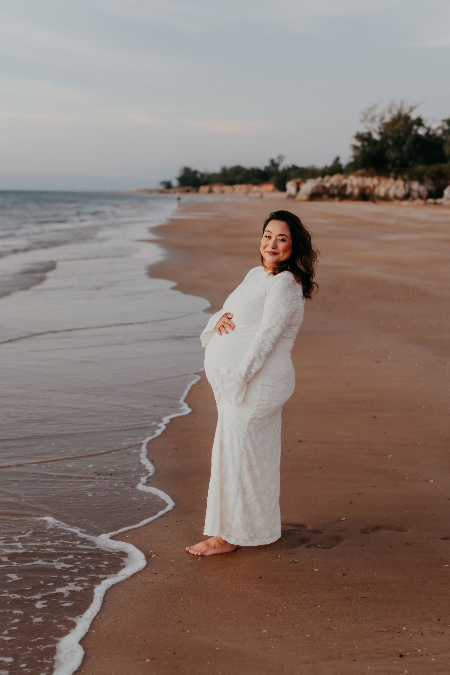 Pregnant woman in white lace dress standing barefoot on a beach at sunset.