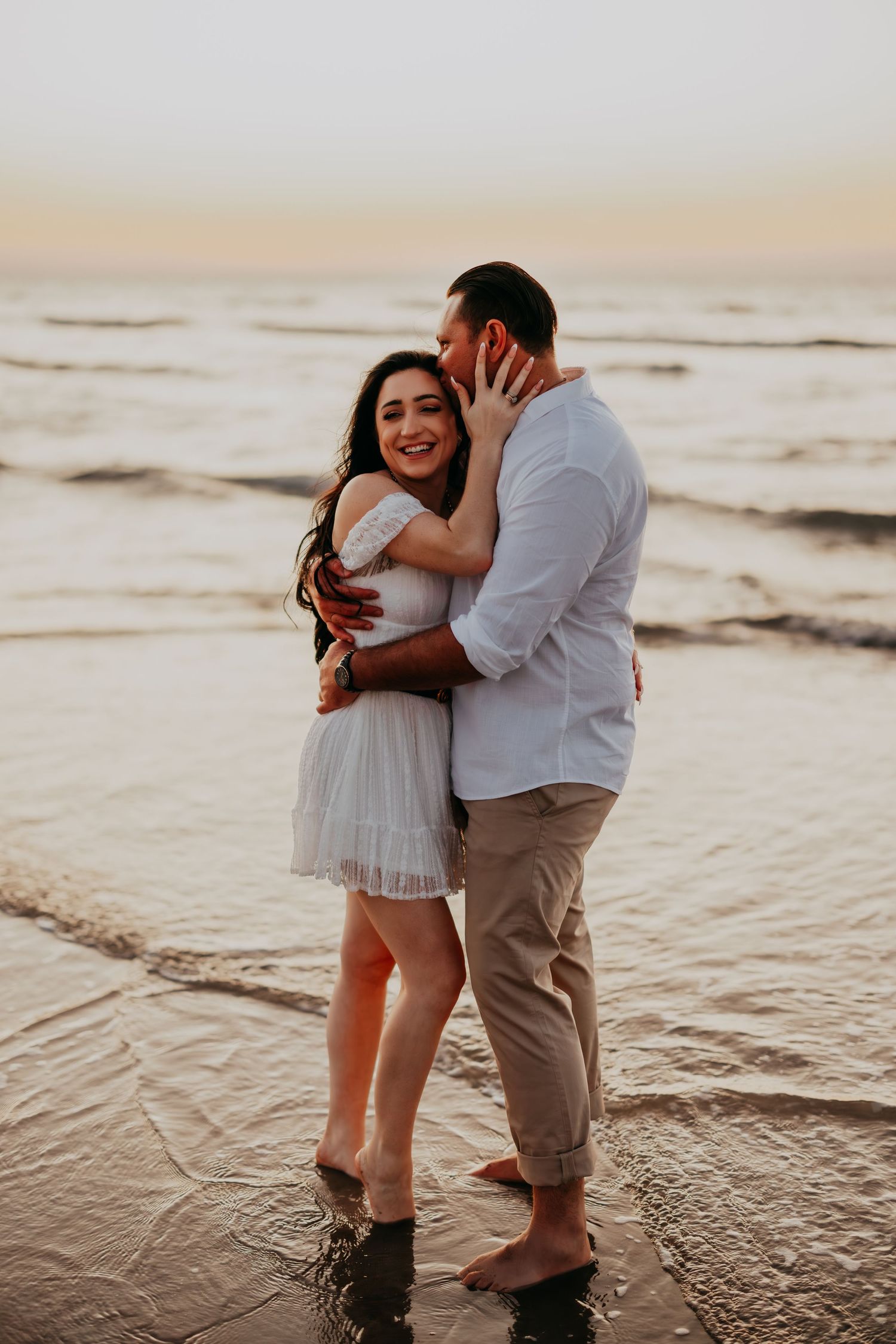 A couple embraces on a wet beach at sunset, the woman in a white dress smiling happily.