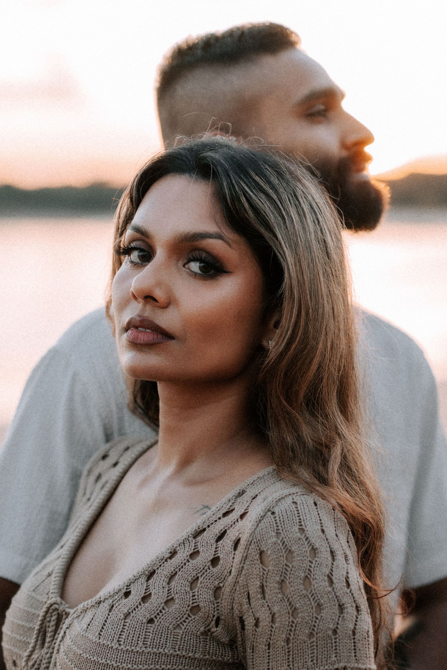 A woman with long hair in a knit top poses with a bearded man at sunset near water.