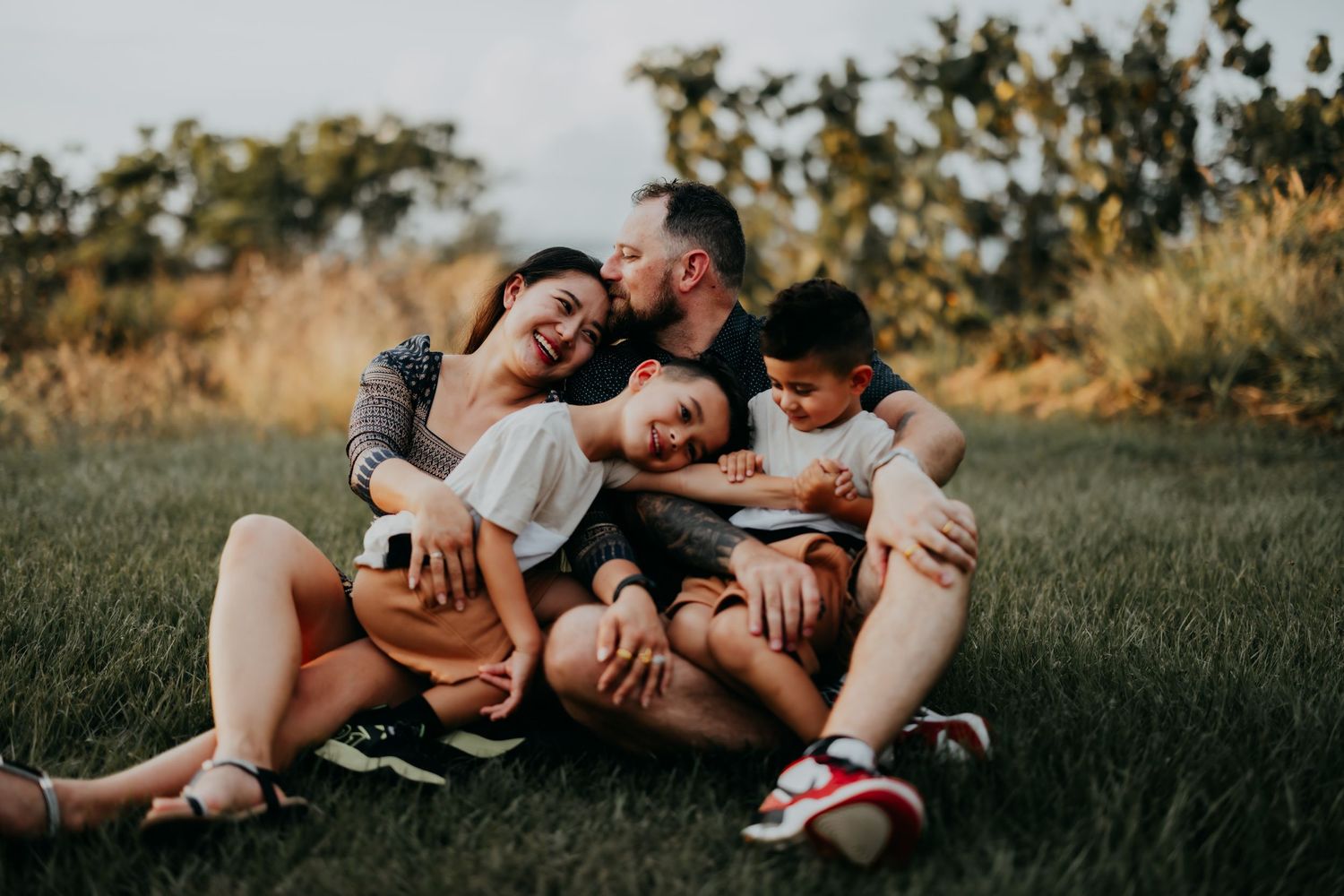 Joyful family of four sitting on green grass, laughing and cuddling together during an outdoor photo session.