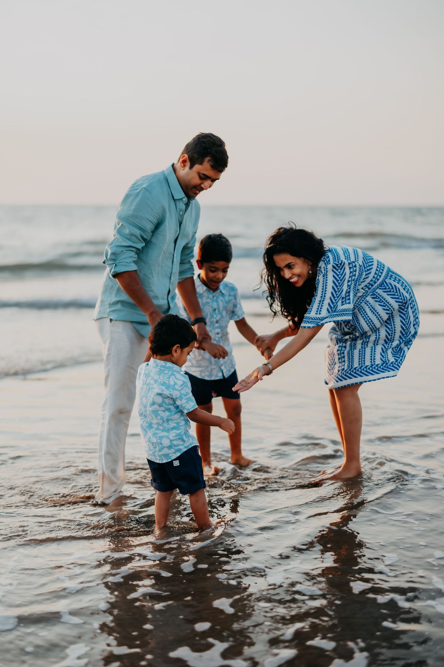 A family of four plays together in shallow ocean waves at sunset on a sandy beach.