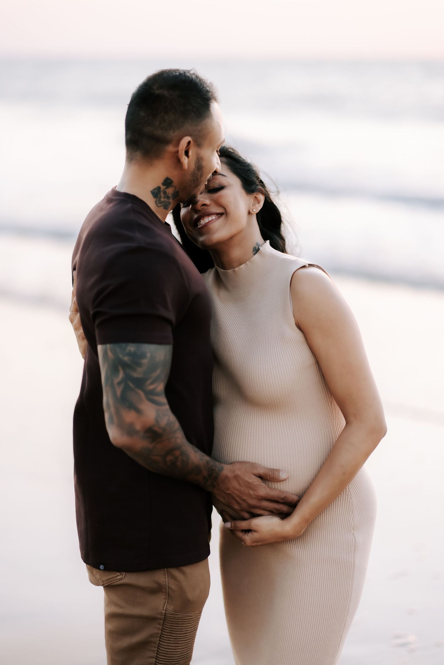 Pregnant couple embracing on a beach at sunset, man with tattoos holding woman's baby bump tenderly.