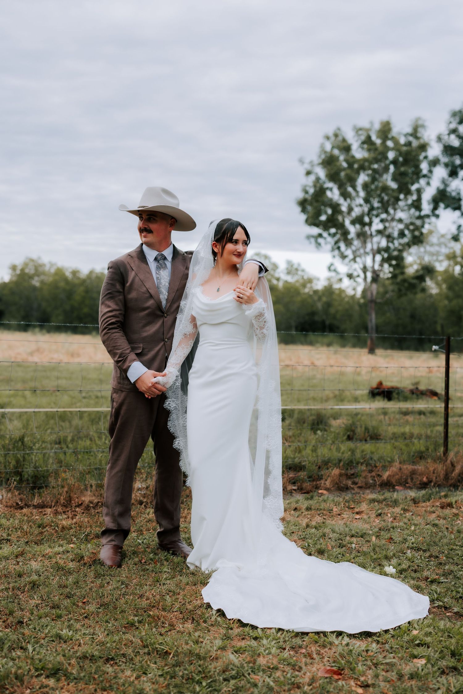 Bride in white lace gown and veil standing with groom in cowboy hat on farm.