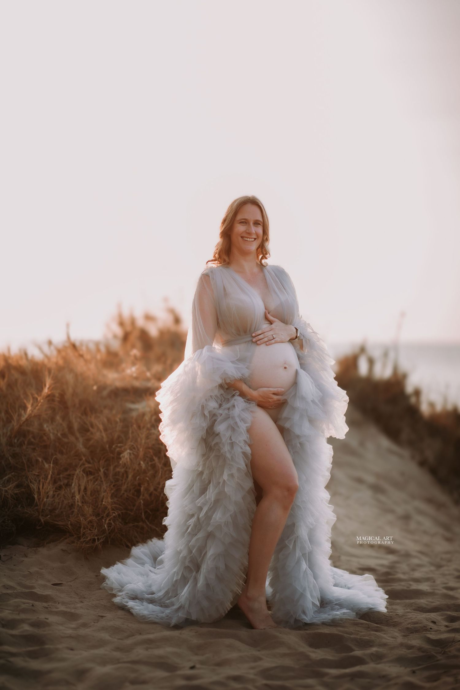 Pregnant woman in white ruffled maternity robe standing barefoot on sandy beach at golden hour.