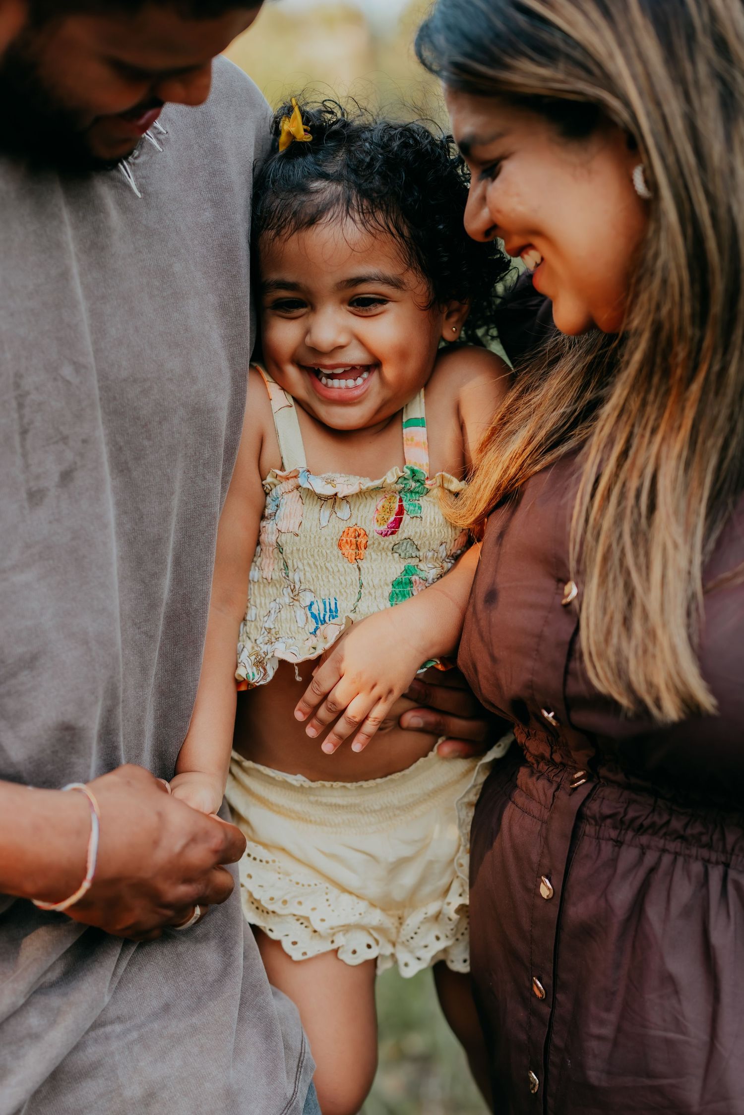 Happy toddler girl laughing joyfully while being held by her smiling parents in an outdoor setting.