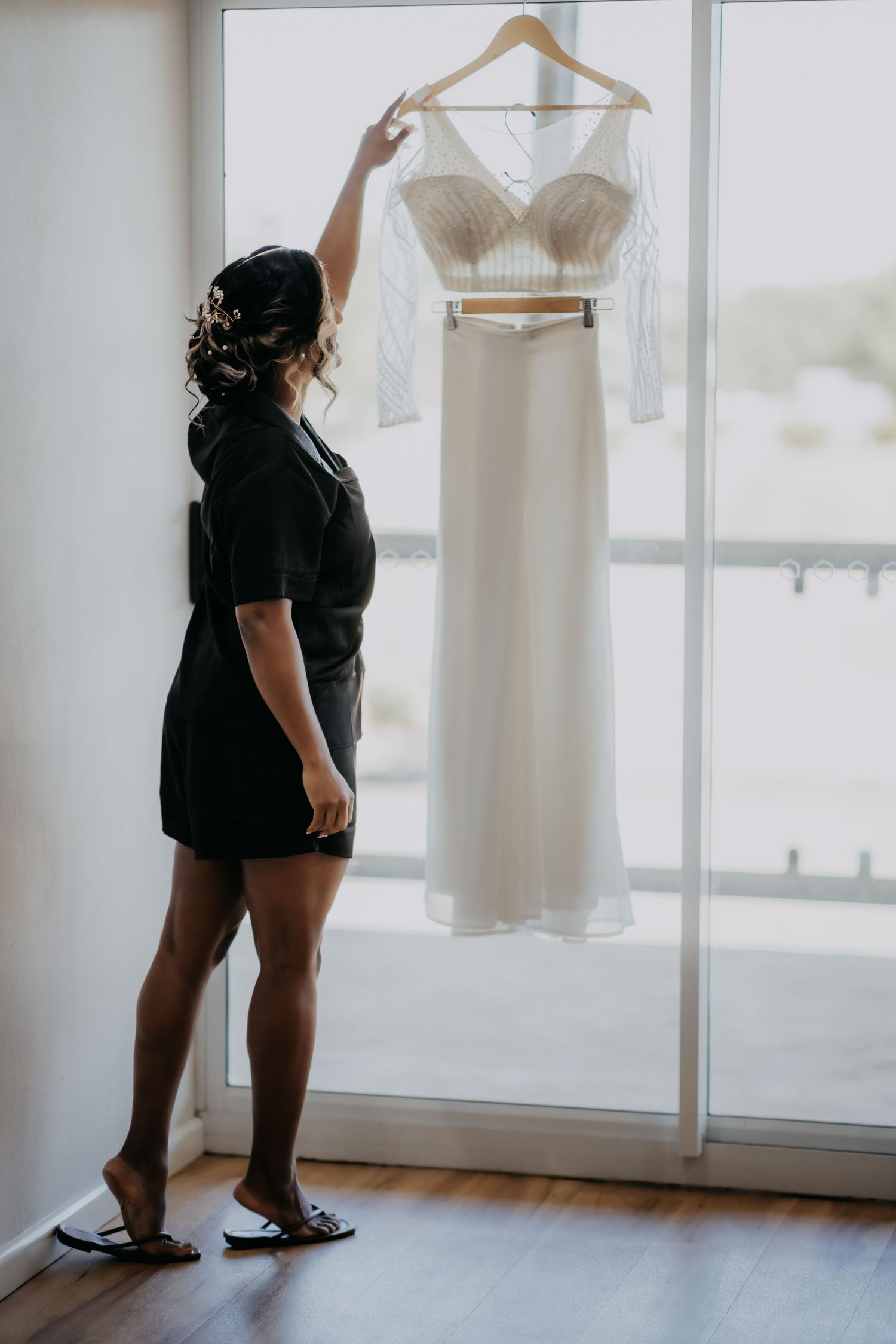 Bride in black robe holding white wedding dress on hanger against bright window light.