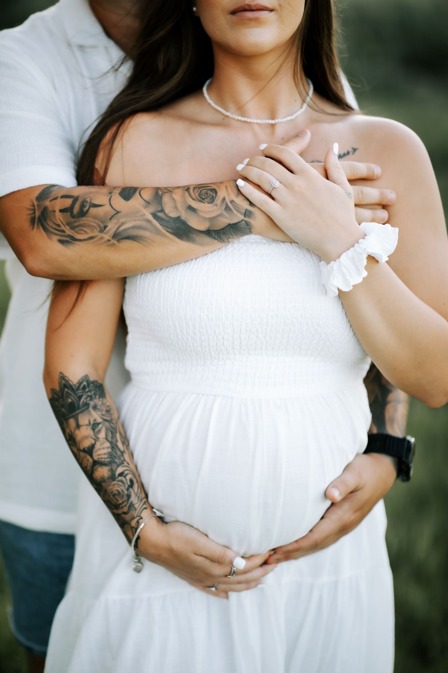 Close-up of tattooed man embracing pregnant woman in white strapless dress outdoors.