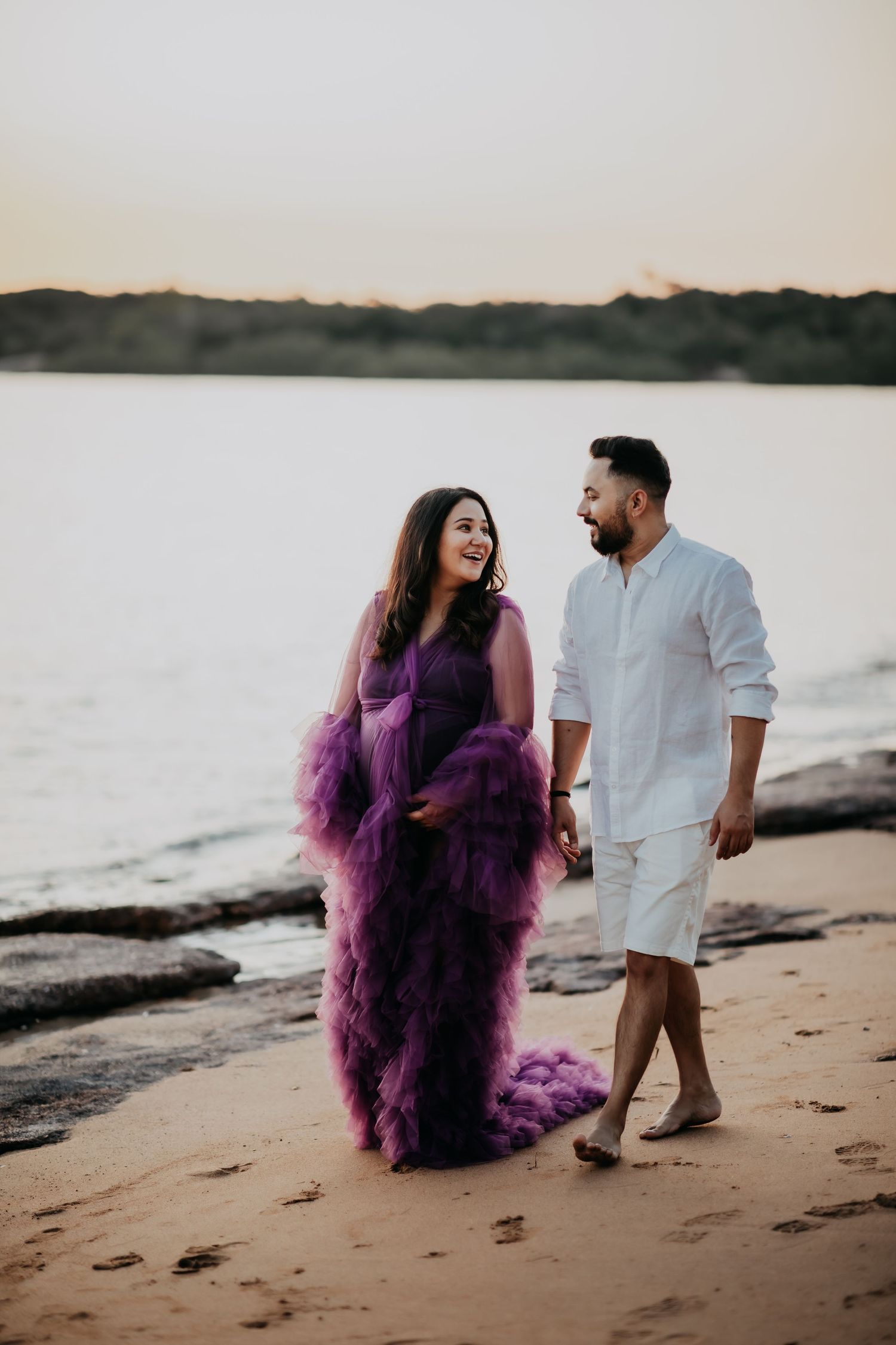 Pregnant woman in purple tulle gown walking hand-in-hand with partner on sandy beach at sunset.
