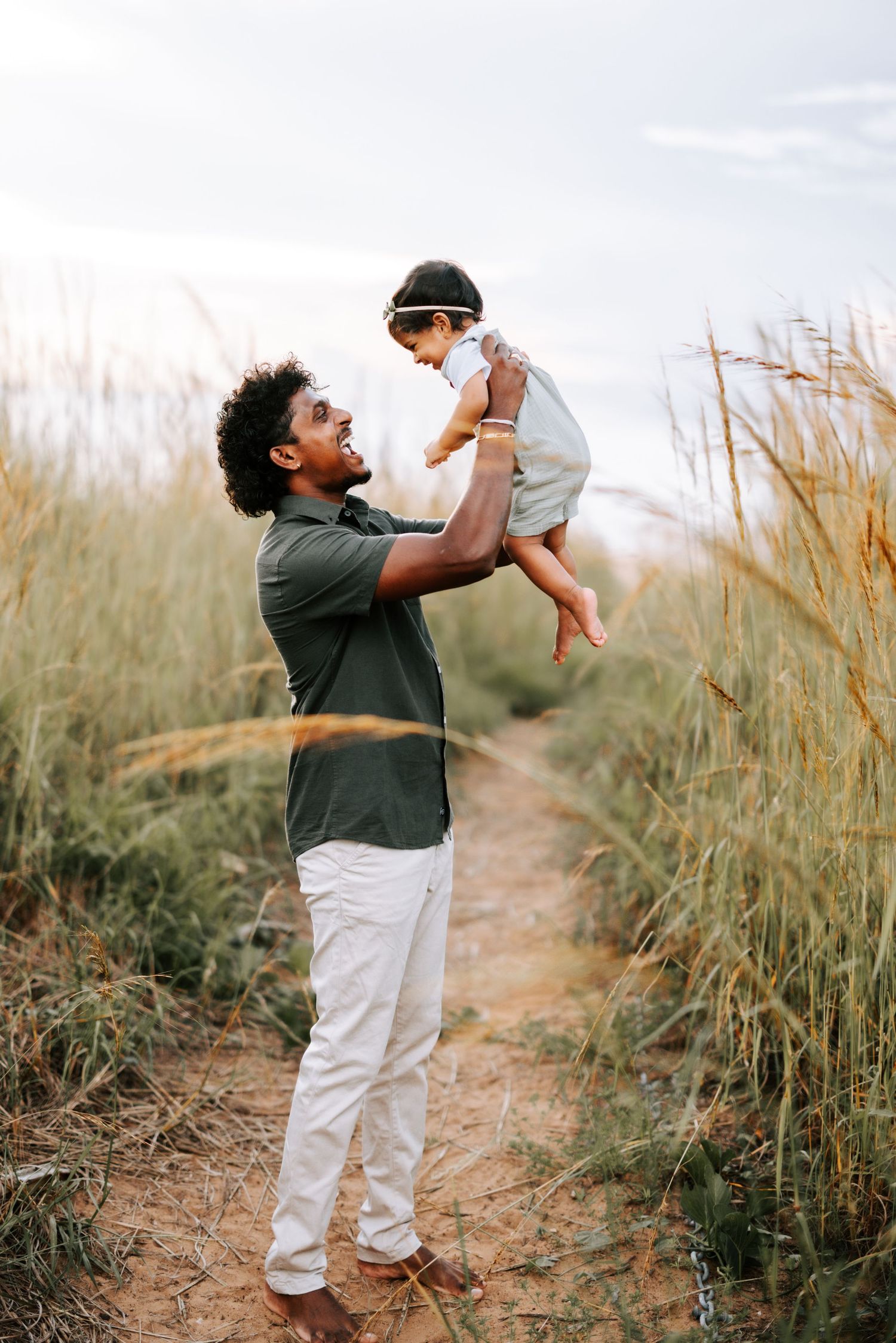 A smiling father joyfully lifts his baby girl high in the air among tall golden beach grasses.