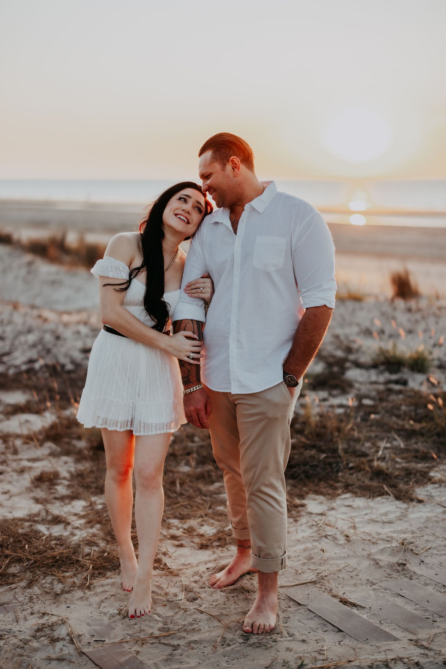 A couple stands together at golden hour on a sandy beach, smiling and embracing warmly.