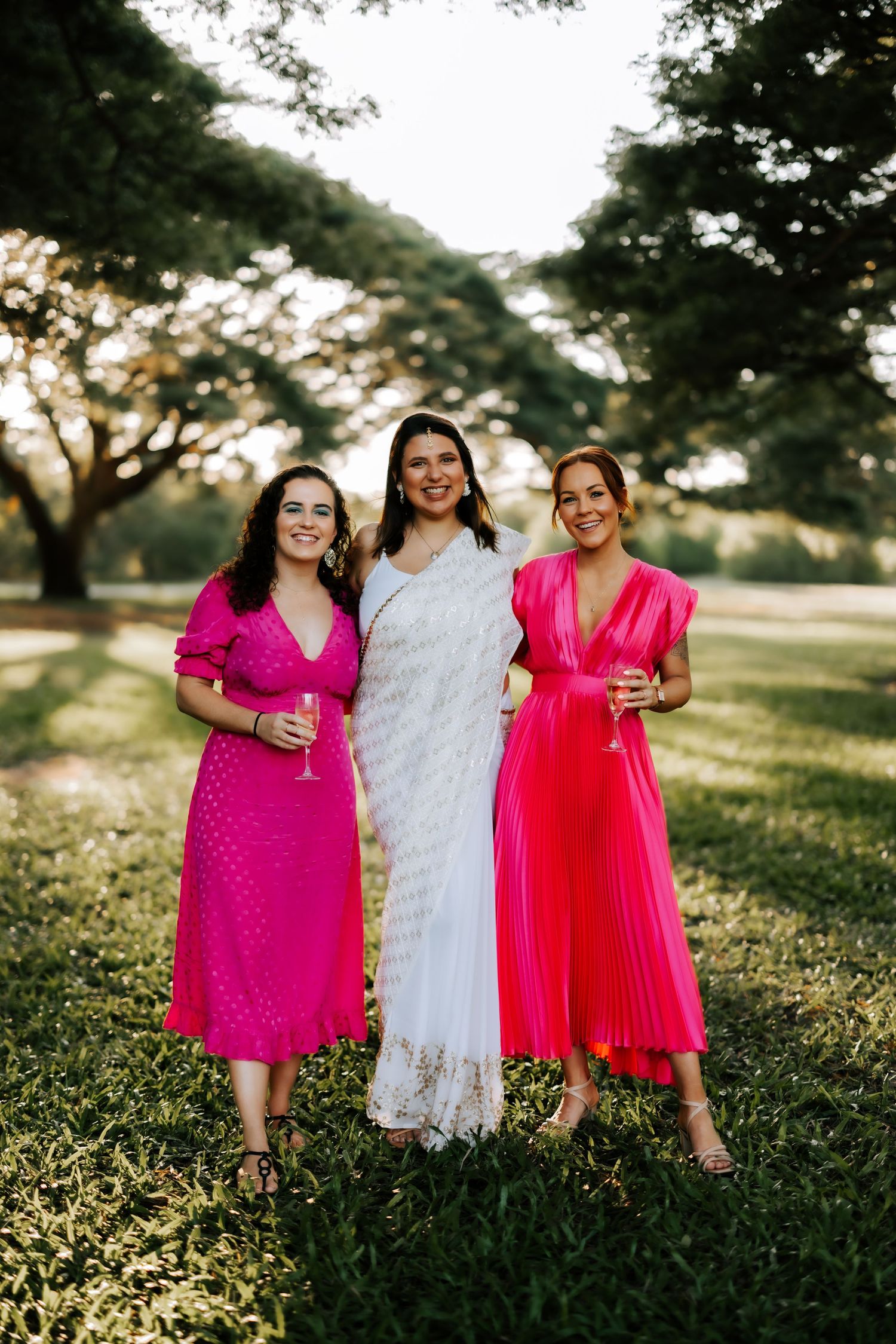 Three smiling women in pink and white saree holding champagne glasses at outdoor wedding celebration.
