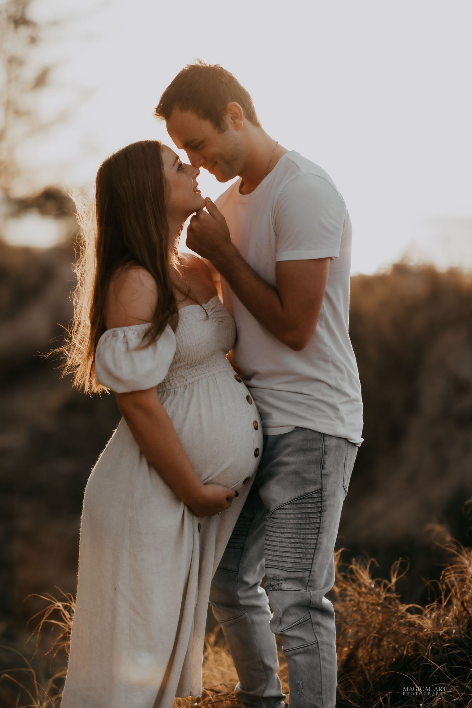 Couple sharing a tender moment outdoors at golden sunset, pregnant woman in white dress.