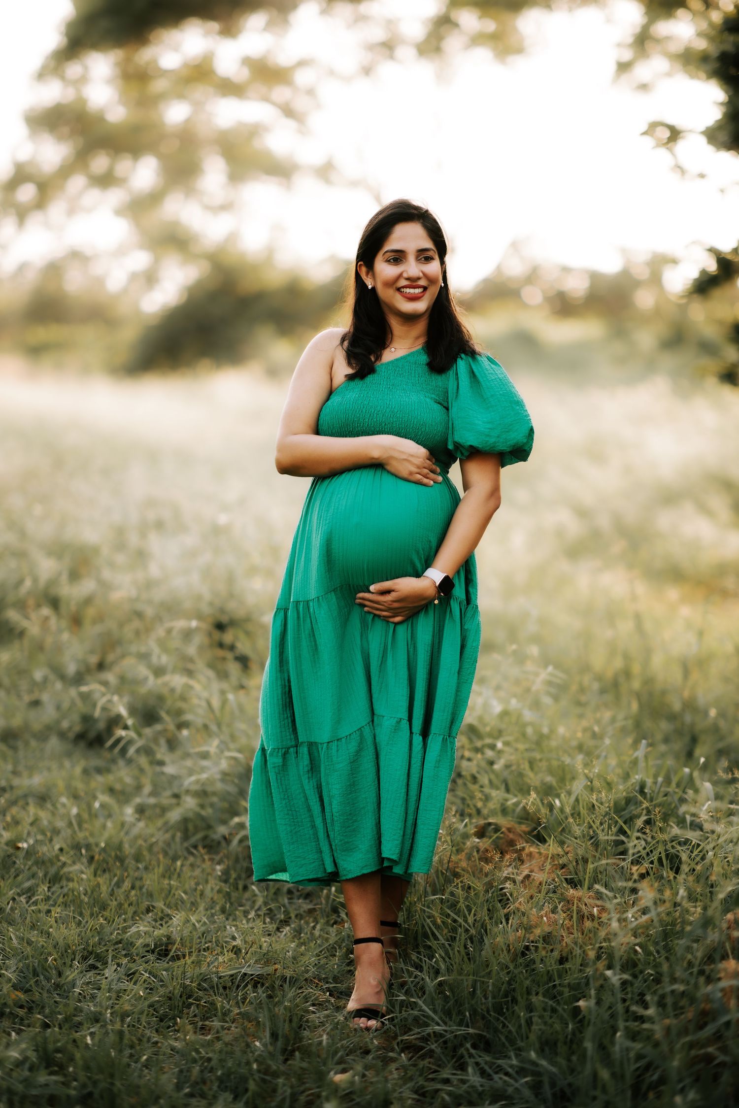 Pregnant woman in green dress holding belly in sunlit field.