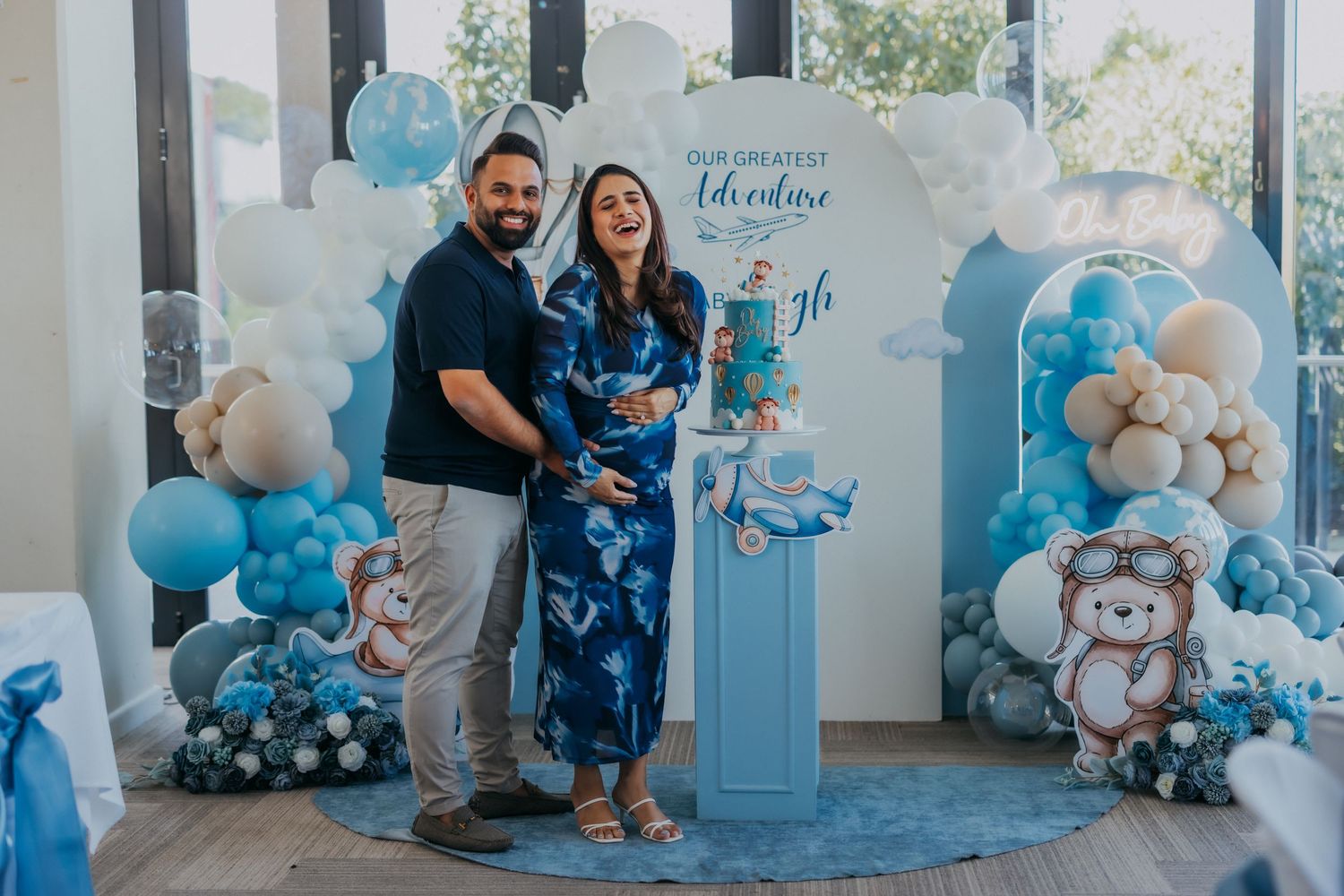 Couple poses at a blue teddy bear baby shower with balloon arch decorations.
