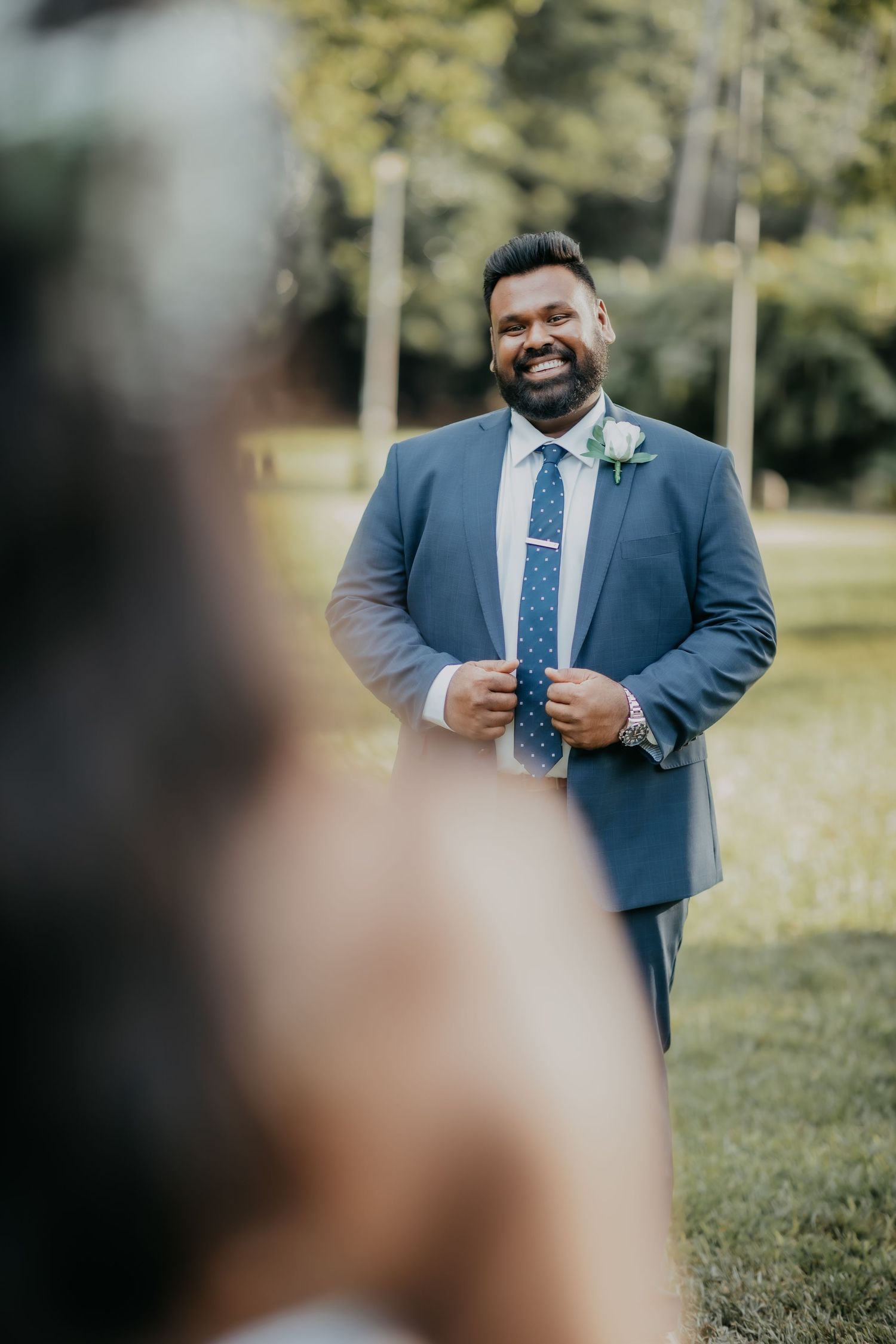 Smiling groom in navy blue suit adjusting tie during outdoor first look wedding moment.