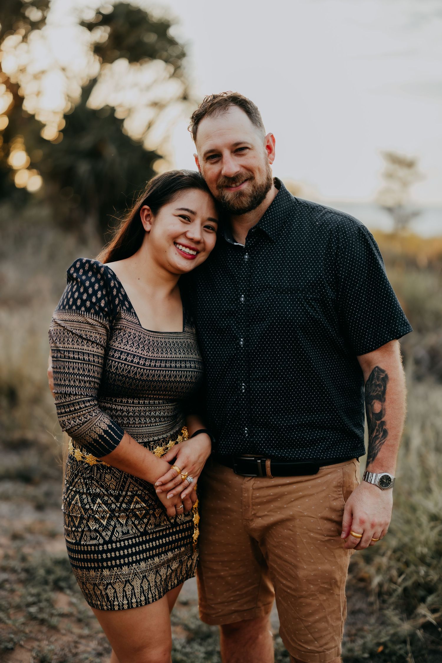 Smiling couple posing together in a sun-kissed outdoor field at golden hour, man with tattoos wearing khaki shorts.